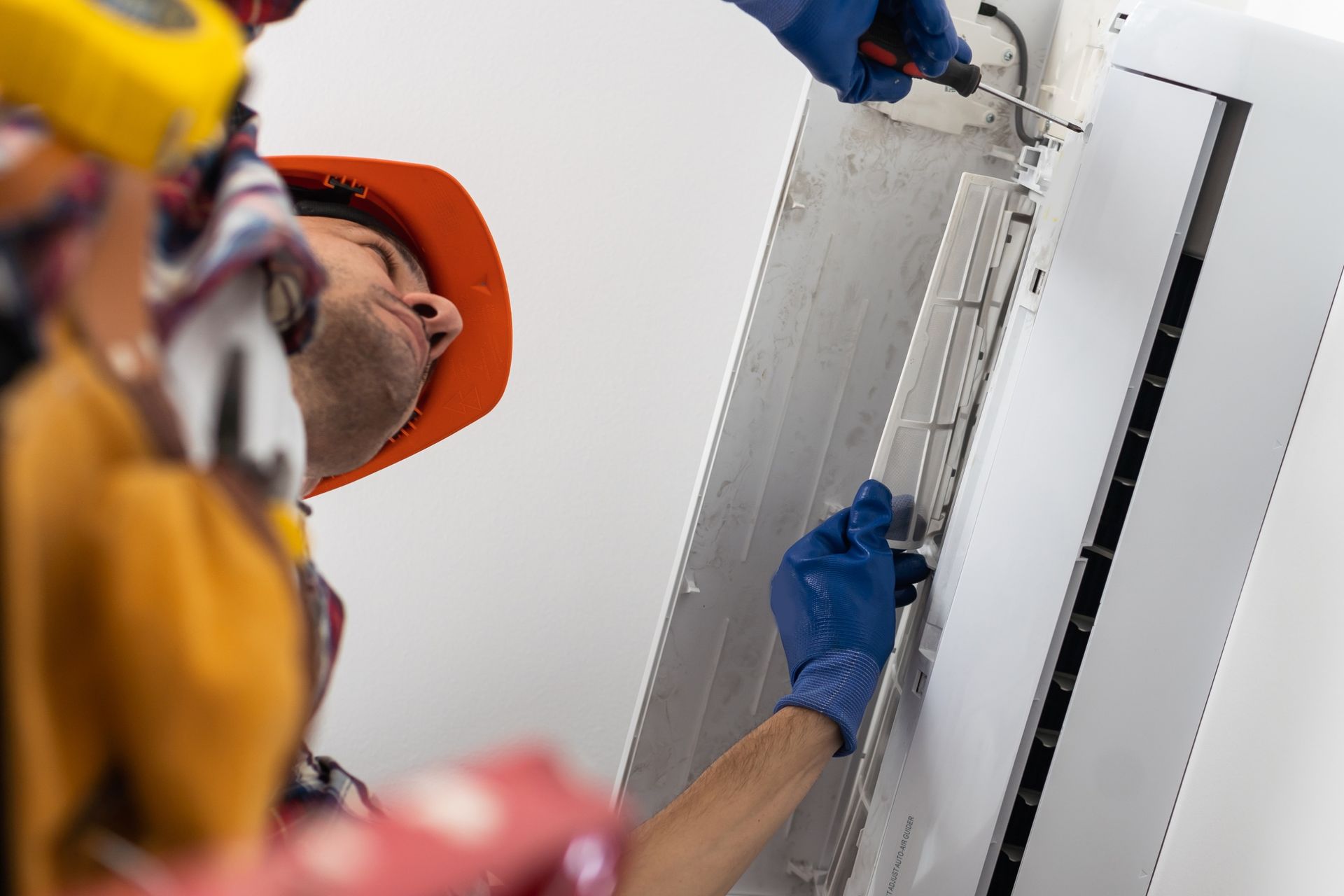 Two workers installing an air conditioning unit; one uses a screwdriver, both wear gloves and hard hats.