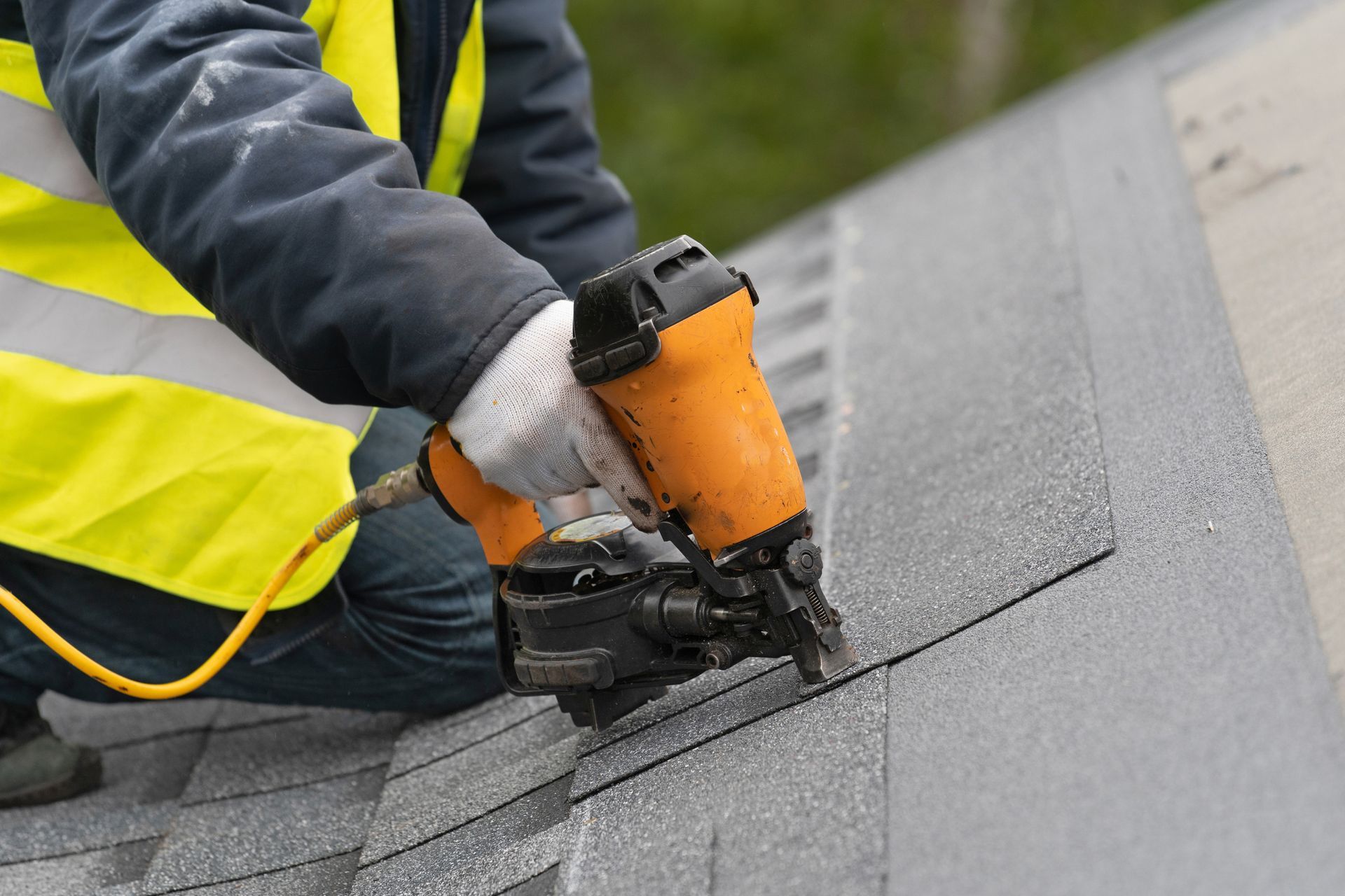 Roofer using a nail gun to secure shingles on a roof. They wear gloves and a safety vest.