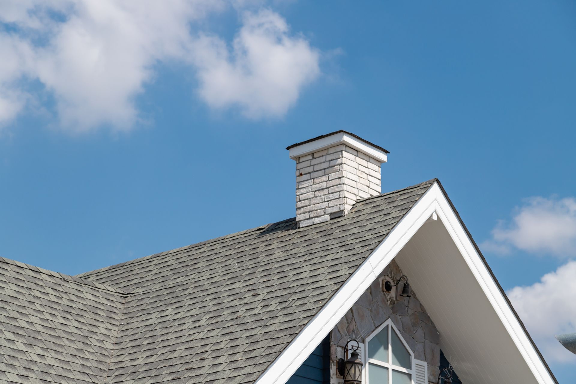 Gray shingled roof with a white brick chimney against a blue sky with clouds.