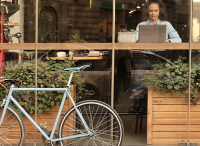 A woman is sitting at a table with a laptop and a bicycle parked in front of a window.