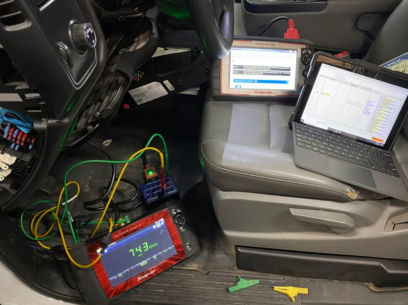 Inside a car, two diagnostic tablets and a laptop connected by cables display software on the floor and seat.