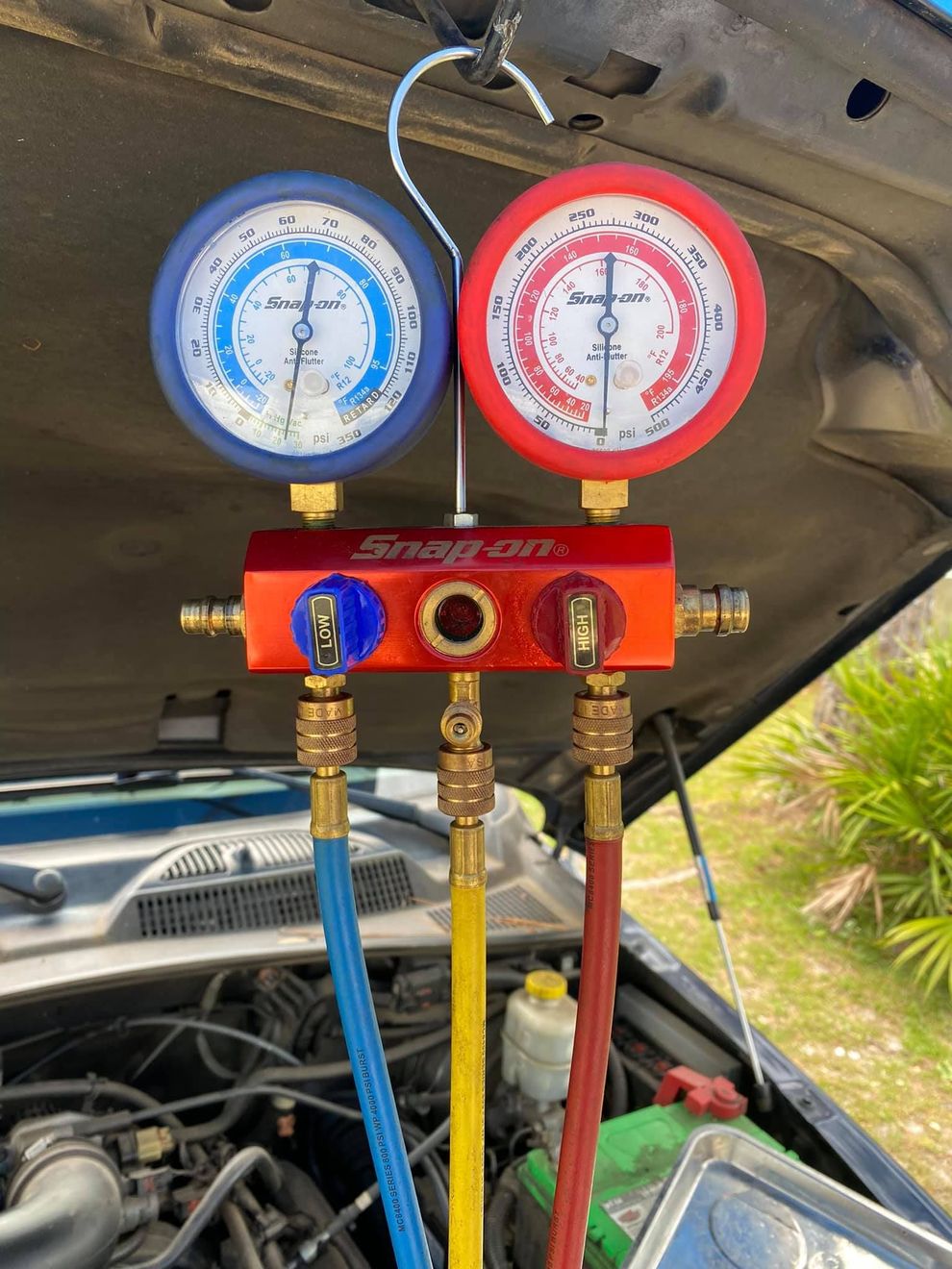 Snap-on AC manifold gauge set hanging from car hood, attached to engine.