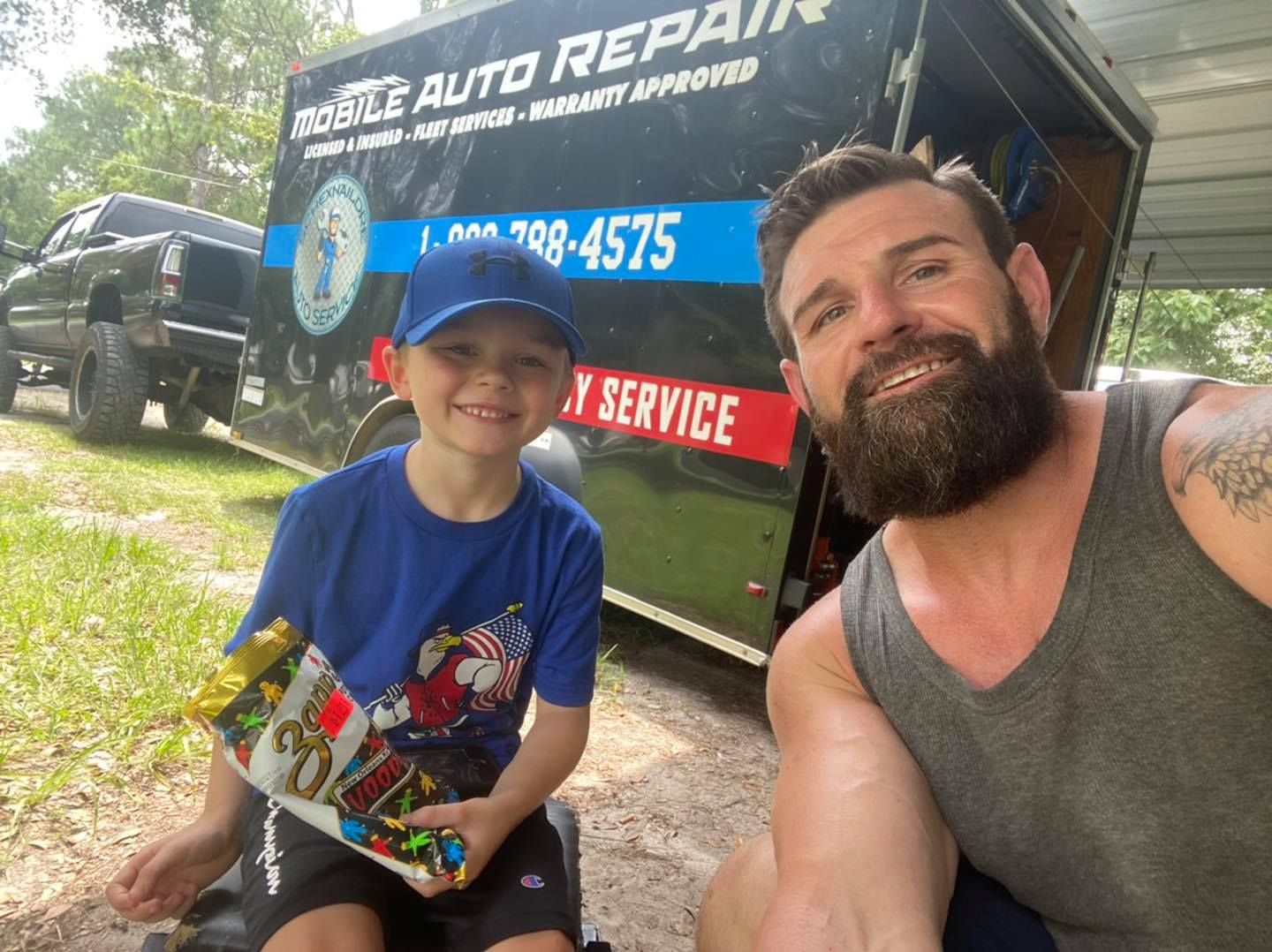 A man and boy smile in front of an auto repair trailer. The boy holds a toy, wearing a blue shirt and cap.