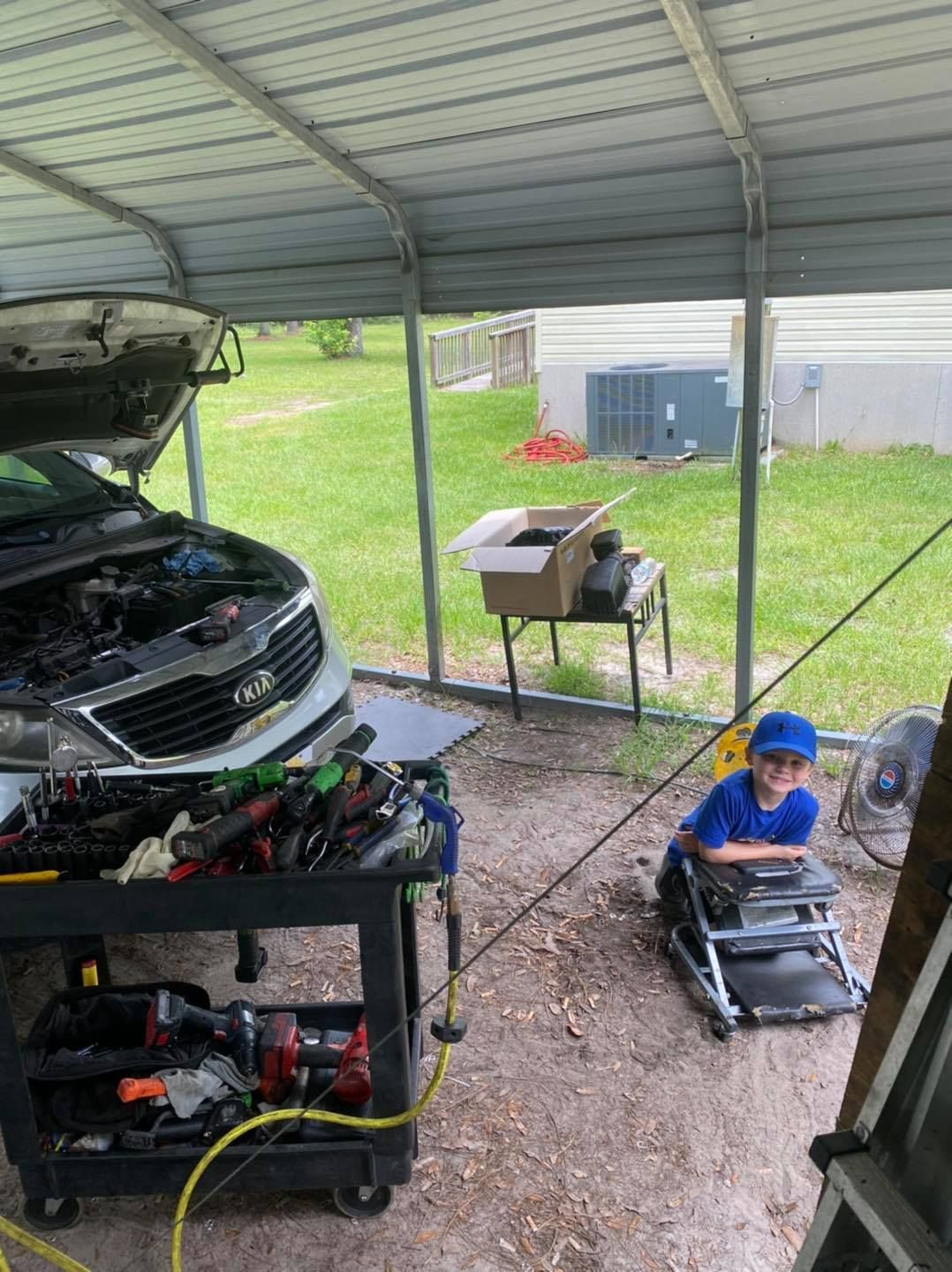 A young person wearing a hat sits on a mechanic's creeper under a carport, near a car with the hood open.