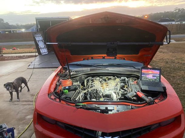 Orange Camaro with open hood, laptop on engine, dog nearby, trailer in background.