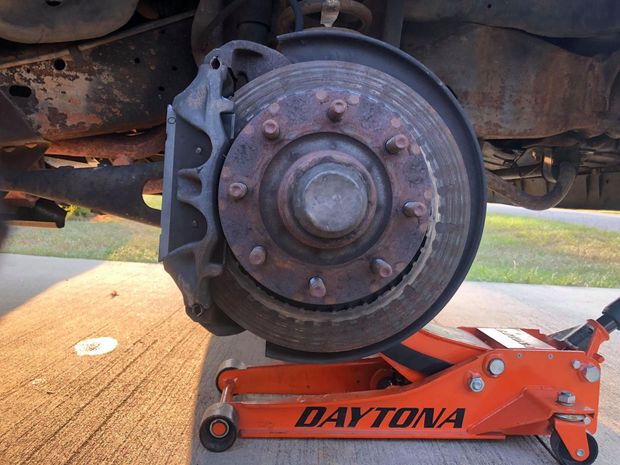 A rusty car brake rotor, caliper, and wheel studs. An orange jack supports the vehicle. Outdoors, daytime.