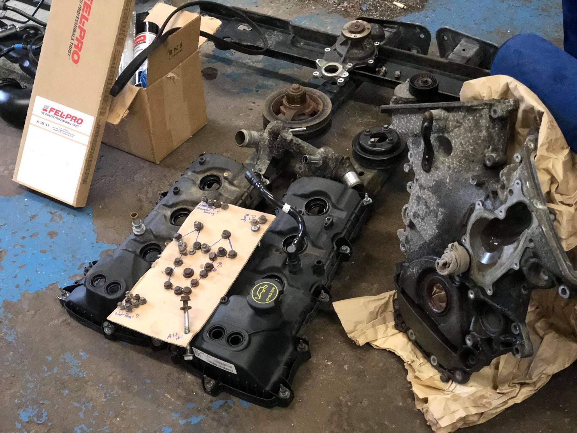 Engine parts scattered on a shop floor: valve covers, engine block, hardware, and boxes, likely for repair.