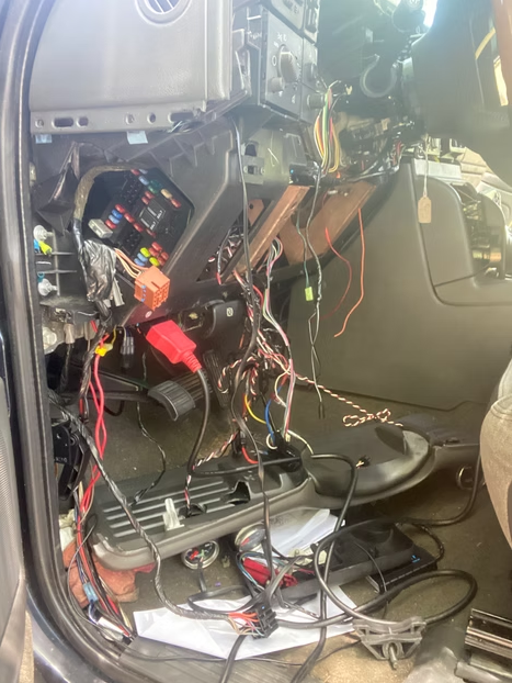 Exposed car dashboard wiring with removed center console and tangled cables inside the passenger footwell