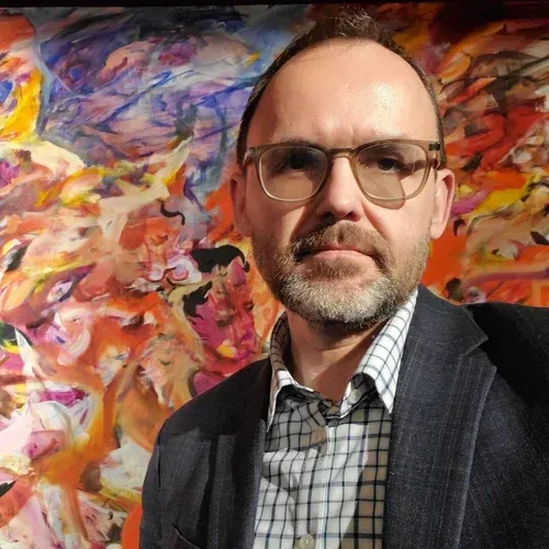 Man in glasses and blazer posing before a vibrant abstract painting backdrop