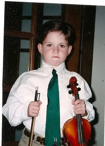 Child in white shirt and green tie holding a violin and bow, standing indoors.