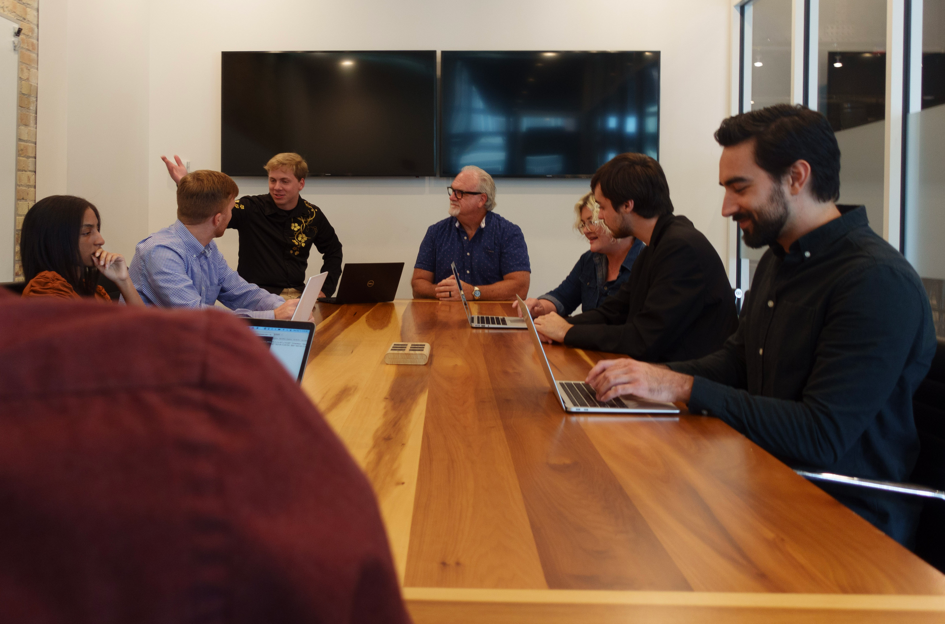 A group of people are sitting around a wooden table with laptops.