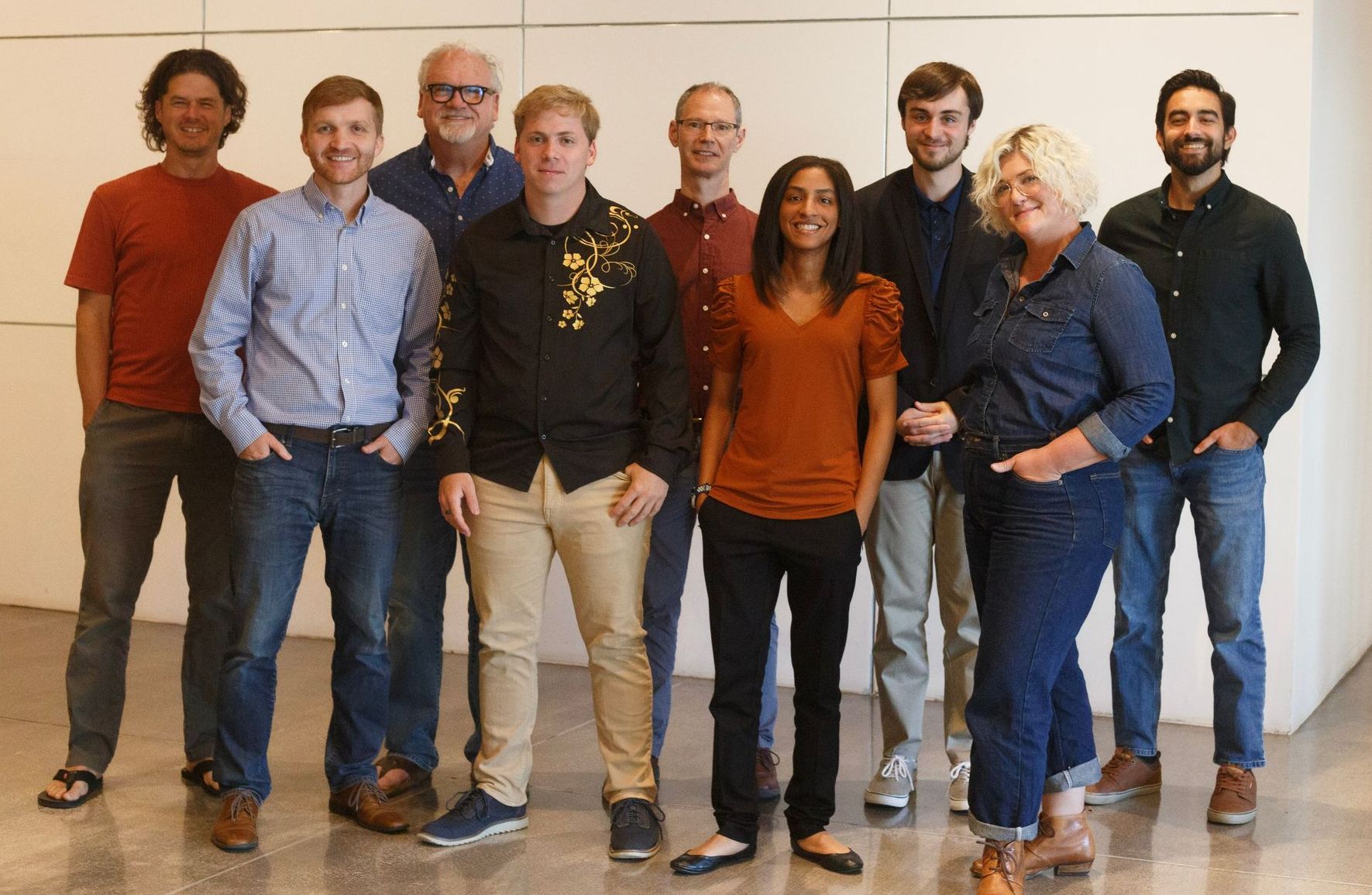 Group of diverse people posing indoors, some smiling. They wear casual clothing.