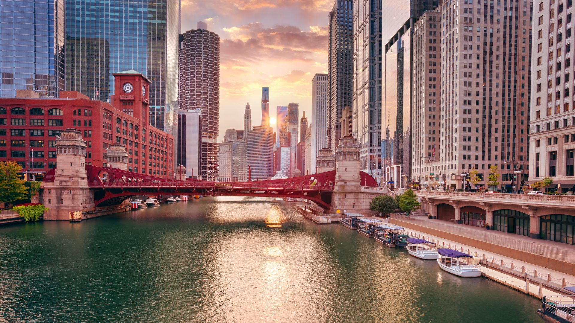 Chicago River at sunset, with bridges, skyscrapers, and boats. Warm light, red bridge, and buildings.