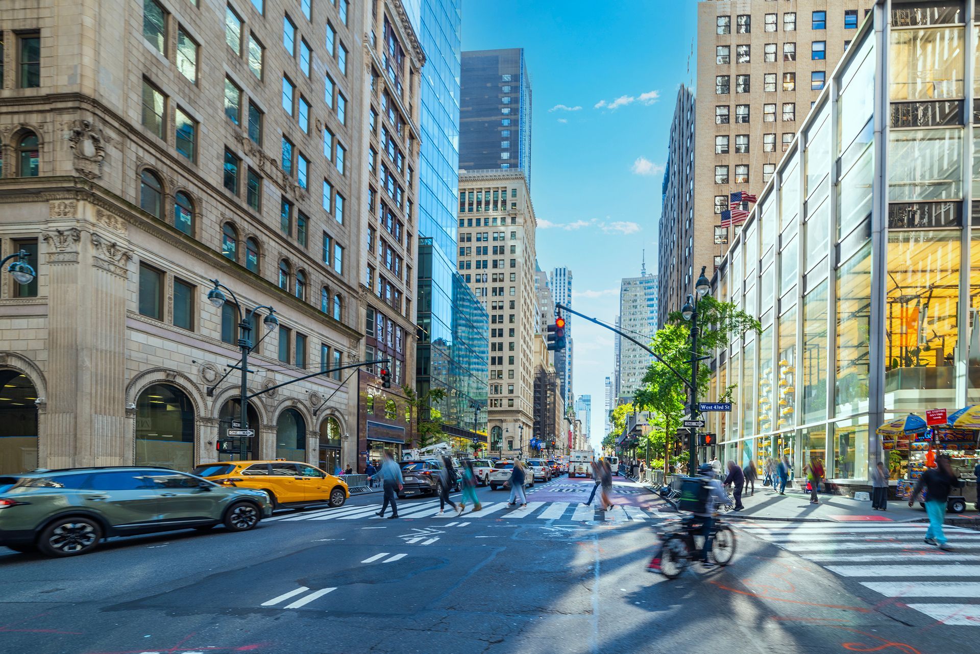 Busy city street with cars, cyclists, and pedestrians between tall buildings on a sunny day