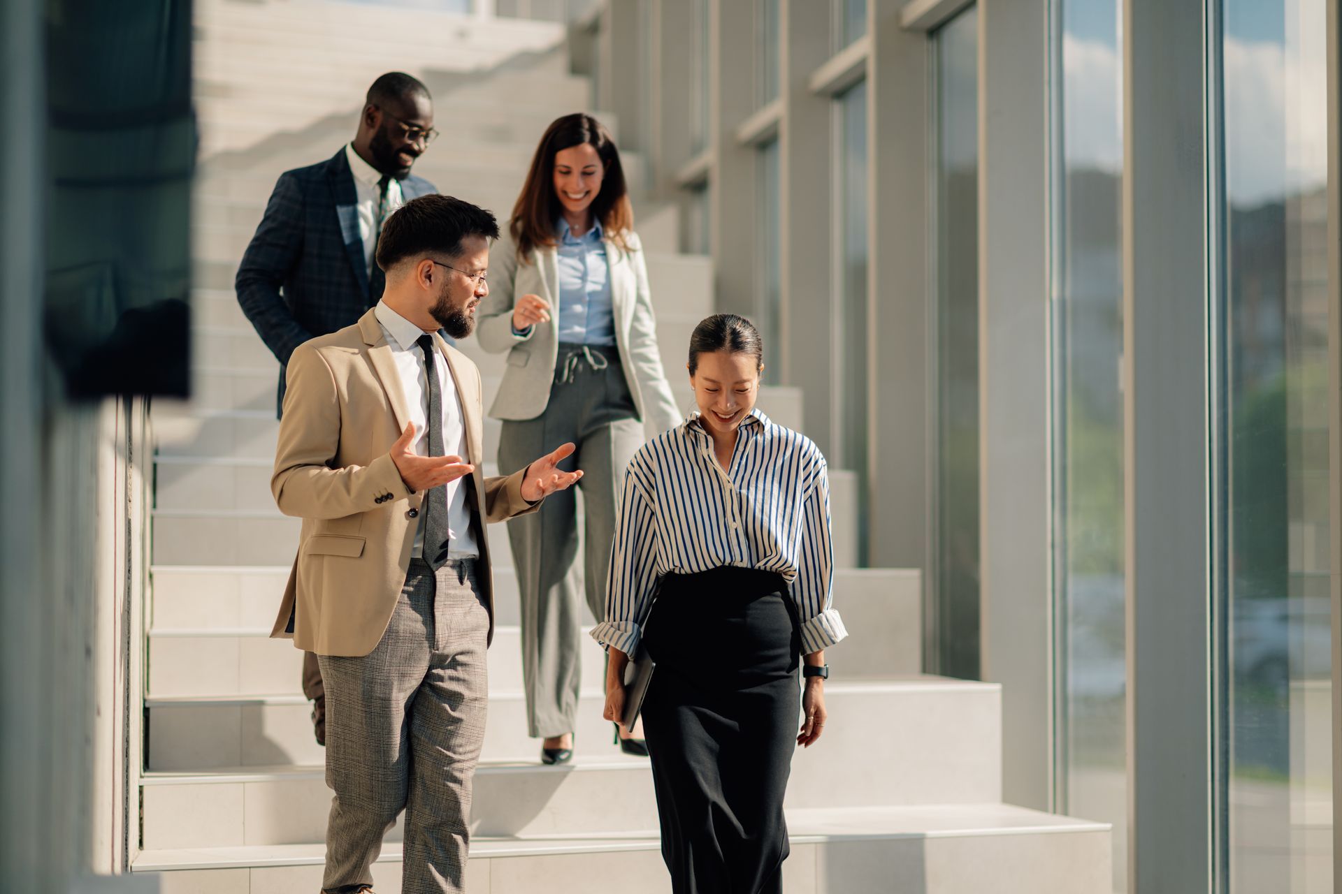 Four coworkers walk down a bright office stairway, one holding a tablet and another speaking with a colleague.