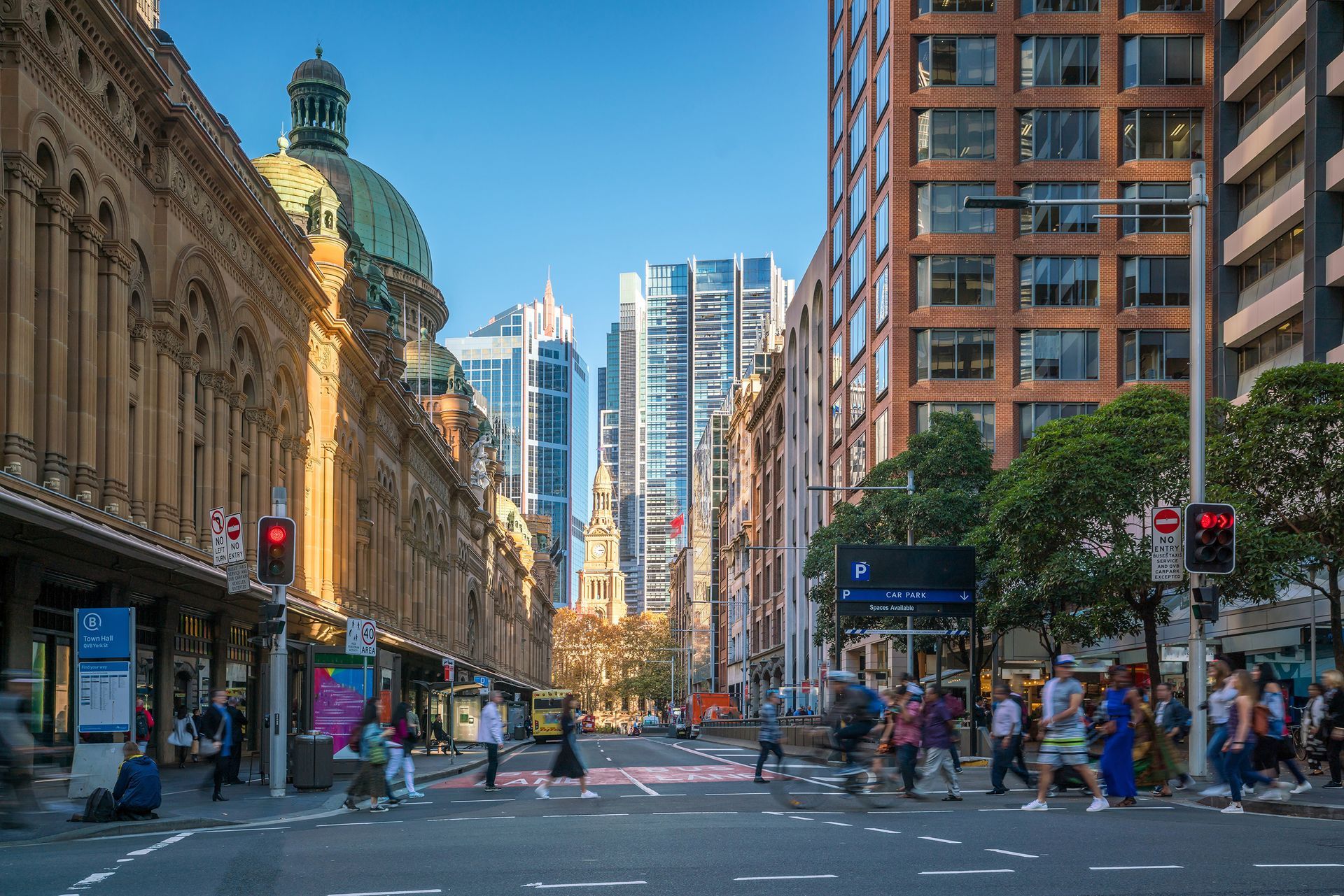 Busy city street with pedestrians, tall buildings, and a domed landmark under a blue sky