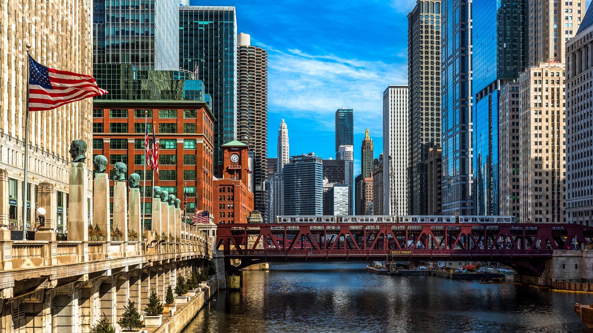 Chicago riverfront with a red bridge, tall downtown buildings, and an American flag along the water