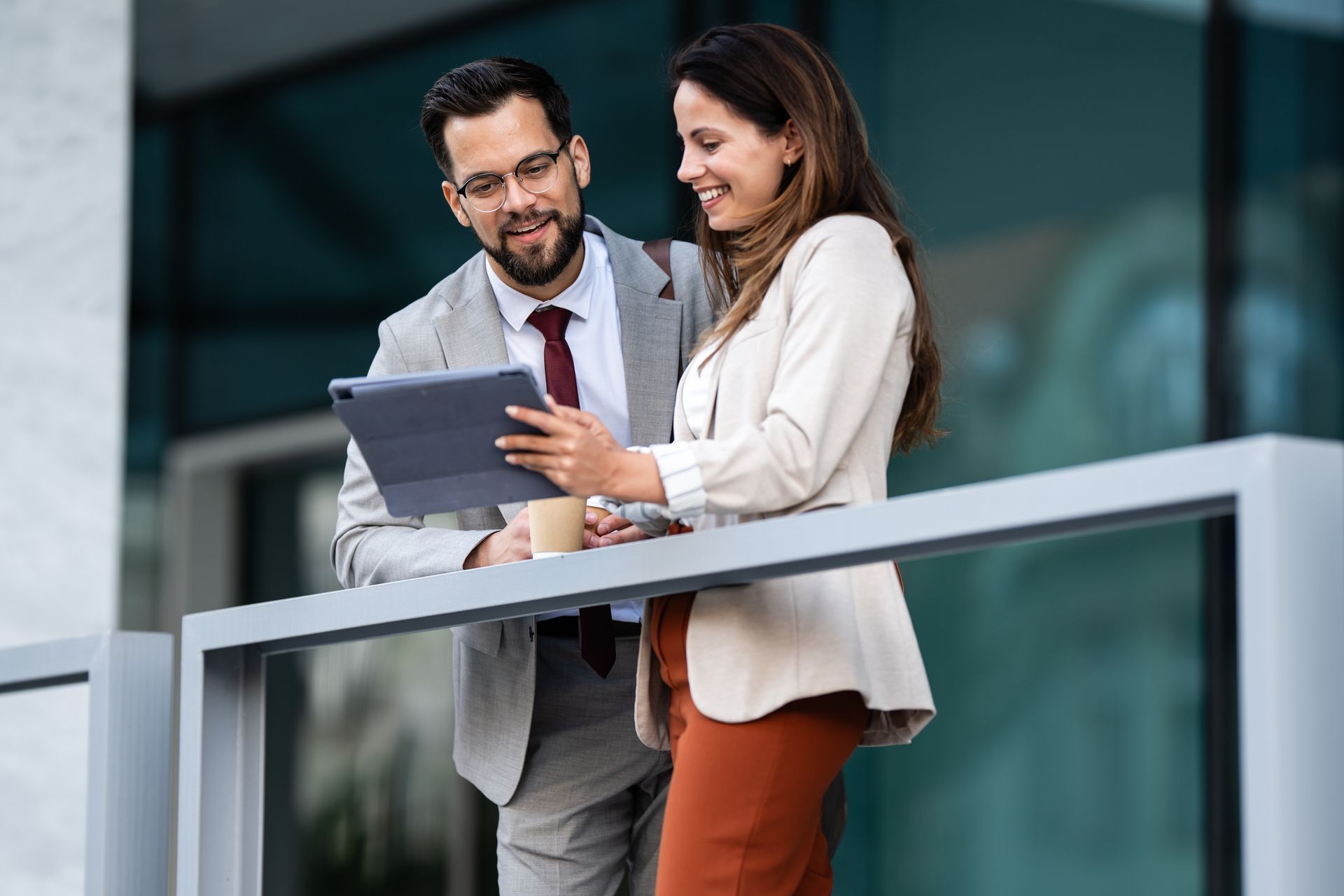 Two coworkers reviewing a tablet and smiling on a balcony outside an office building