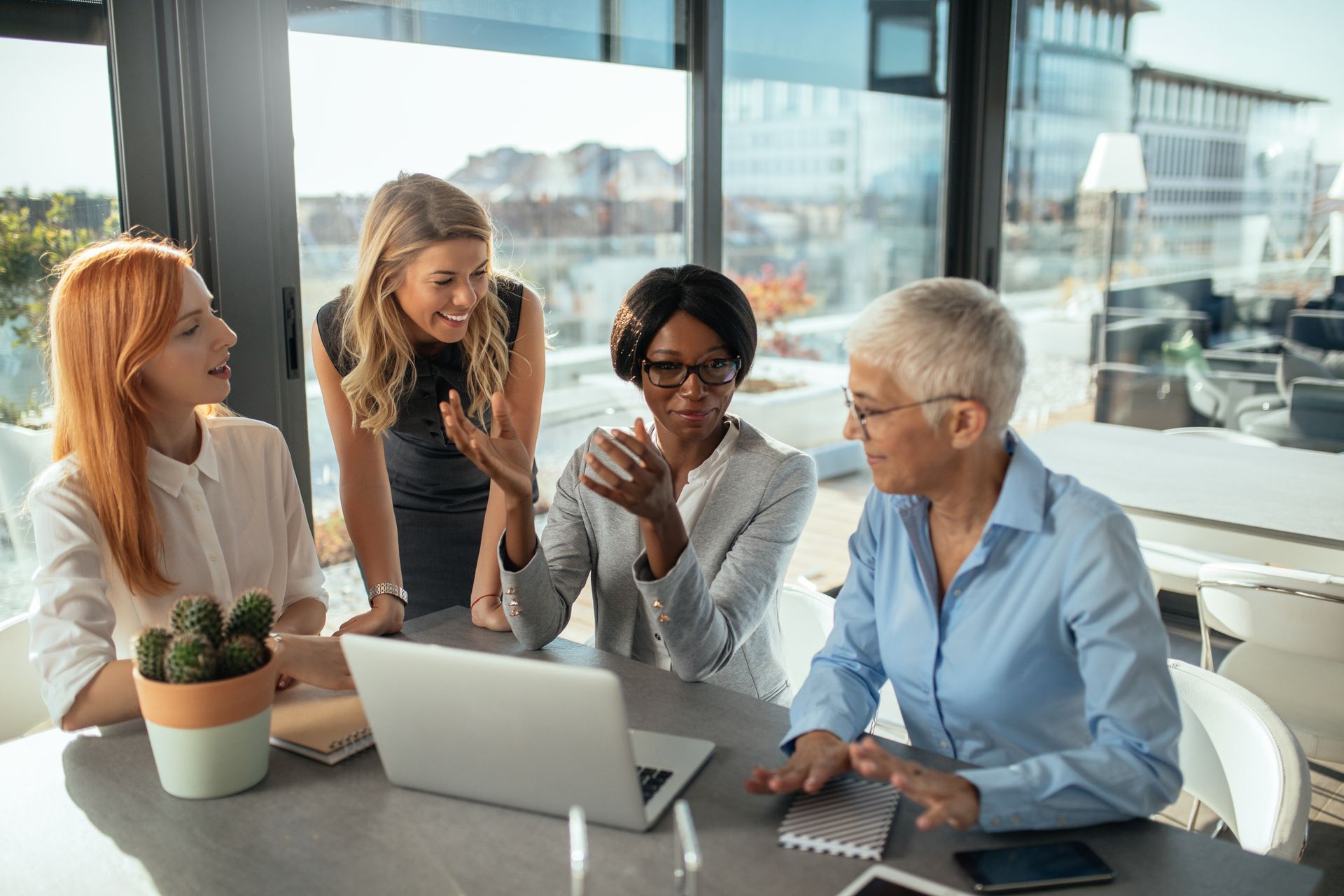 Four coworkers in a bright office meeting around a laptop, one standing and speaking while others listen and smile