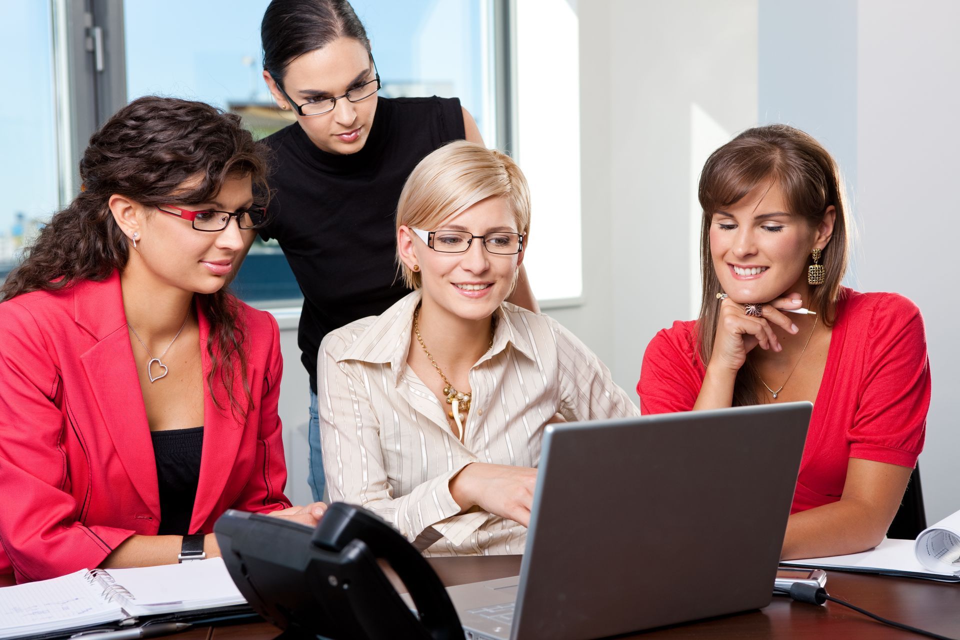 Four coworkers gathered around a laptop, smiling and discussing work in an office.