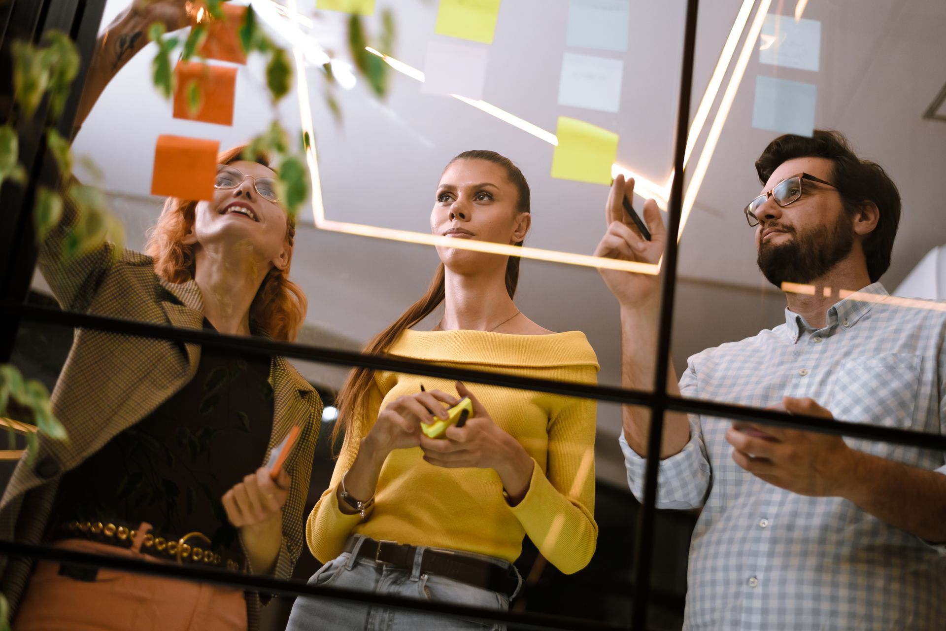 Three coworkers place colorful sticky notes on a glass board in a bright office.