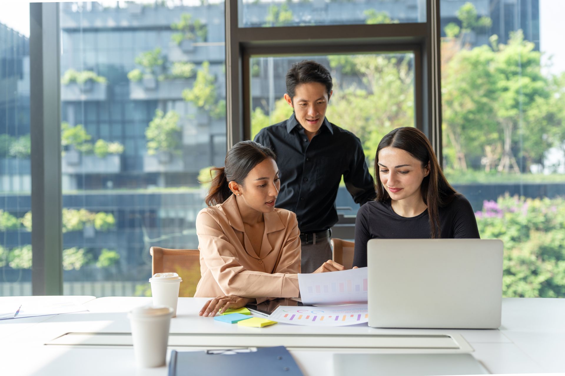 Three coworkers reviewing documents at a laptop in a bright office