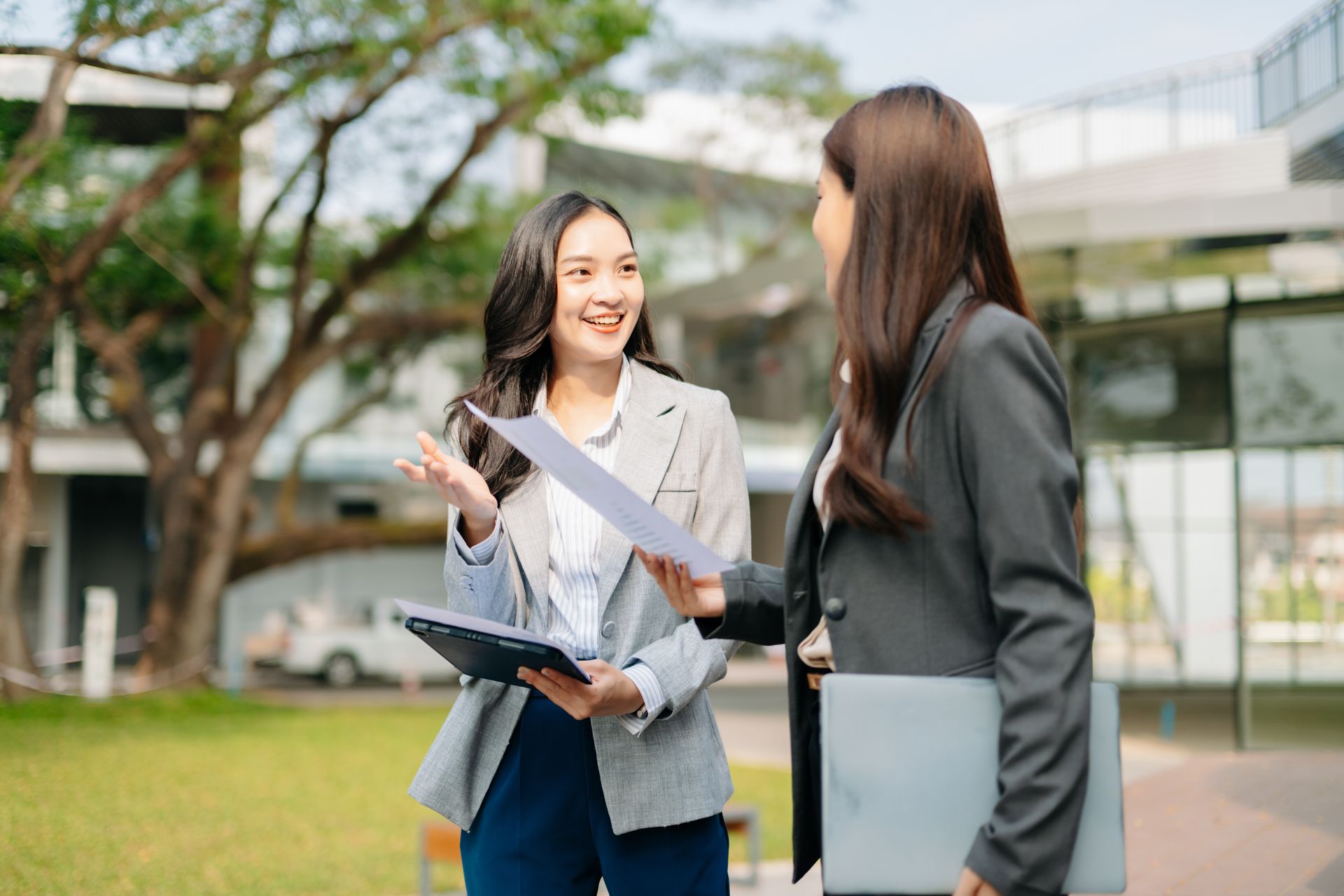Two people talking and smiling outdoors, holding papers and a laptop on a campus lawn