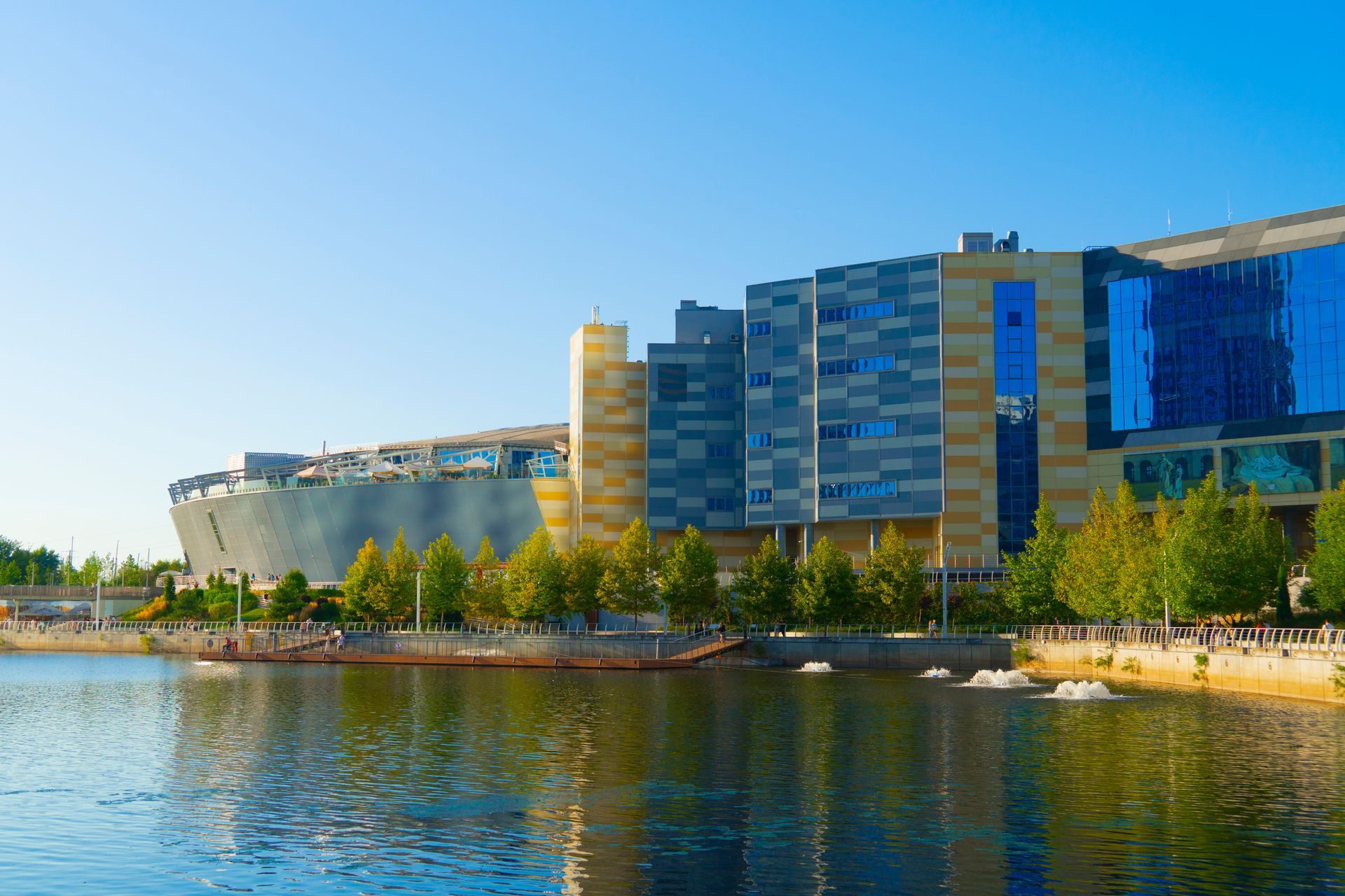 Modern waterfront hotel and adjacent building reflected in calm water under a clear blue sky.