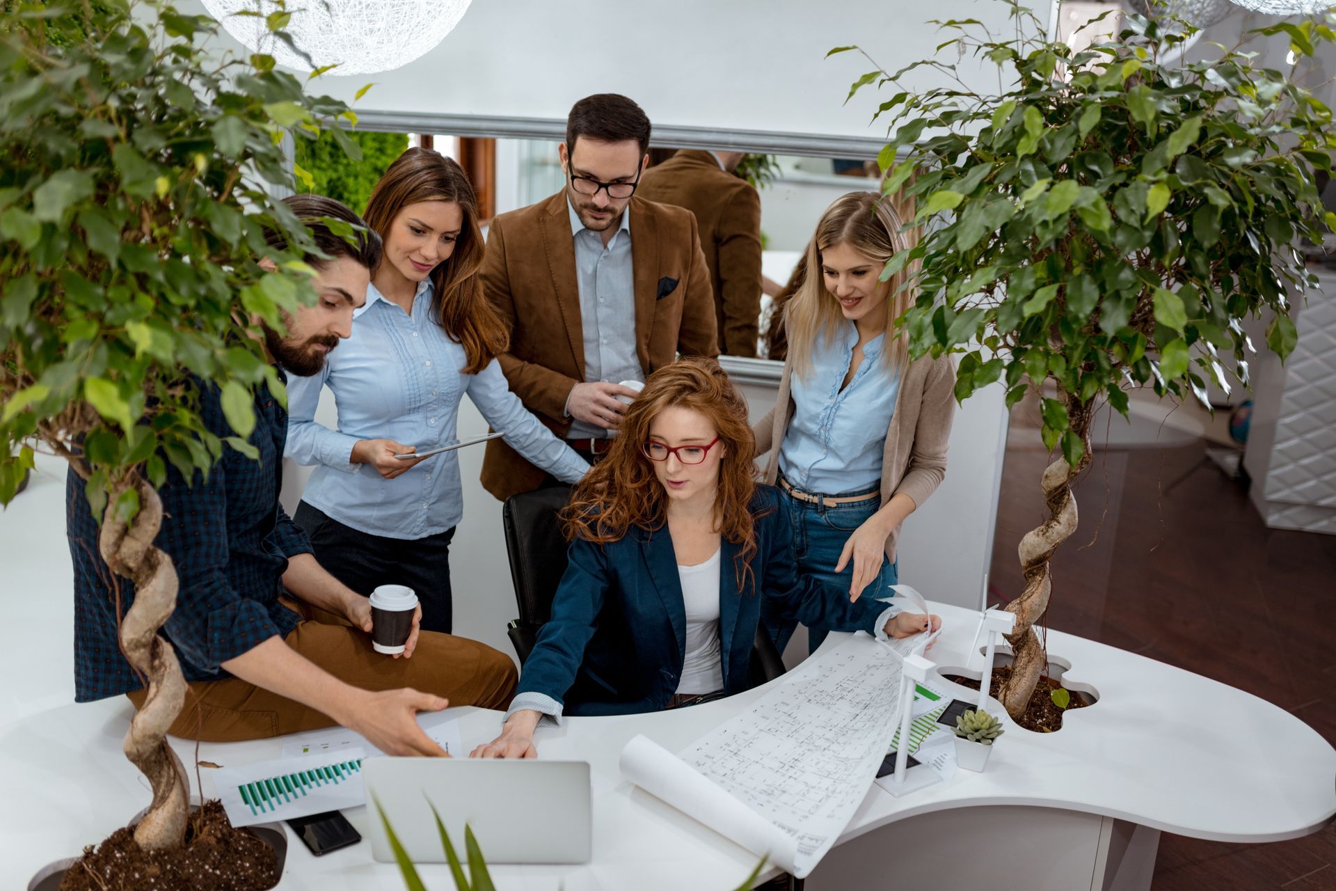 Group of people around a desk looking at a laptop and blueprints in an office with trees.