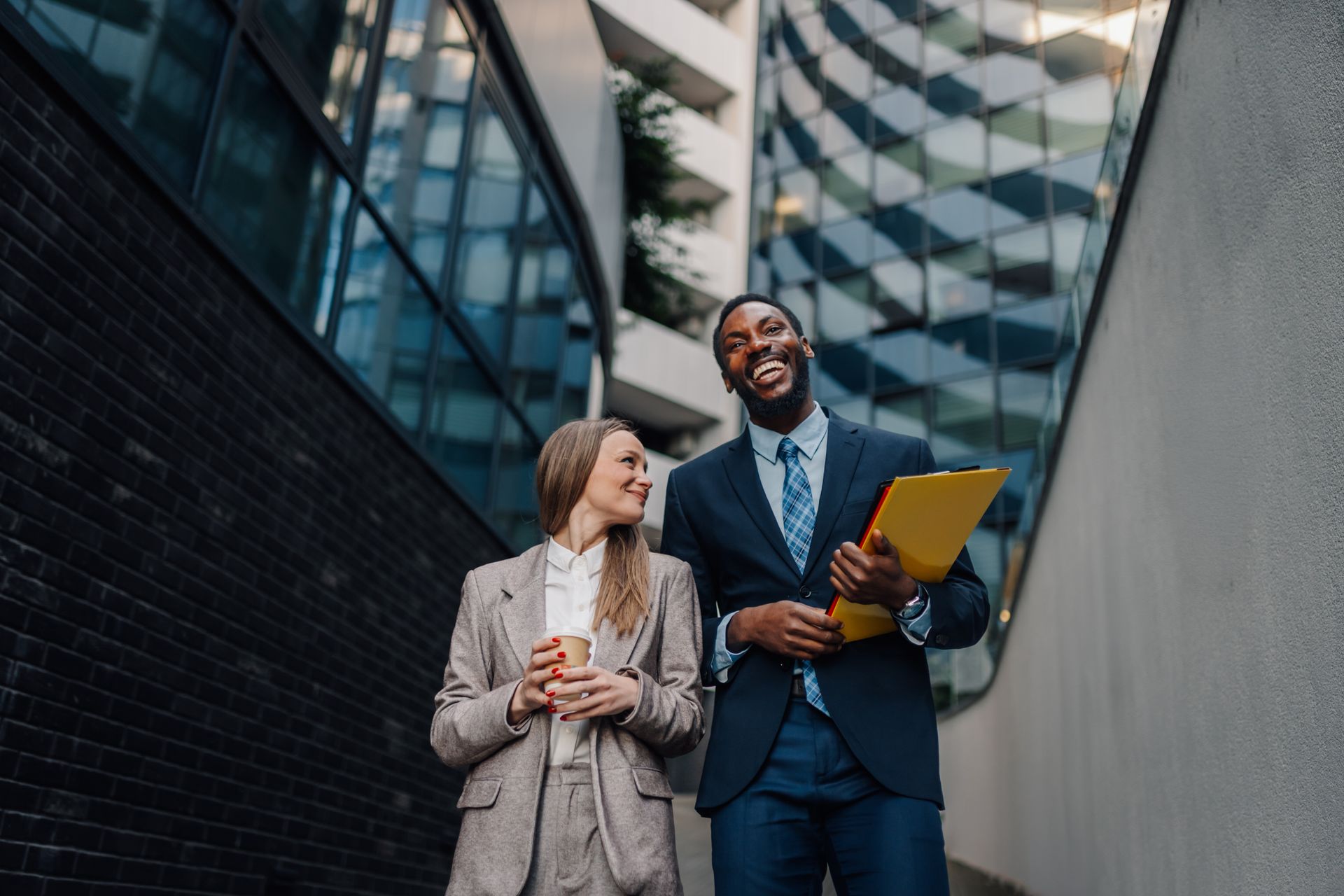 Two professionals walking outdoors between modern buildings, smiling and reviewing a yellow folder