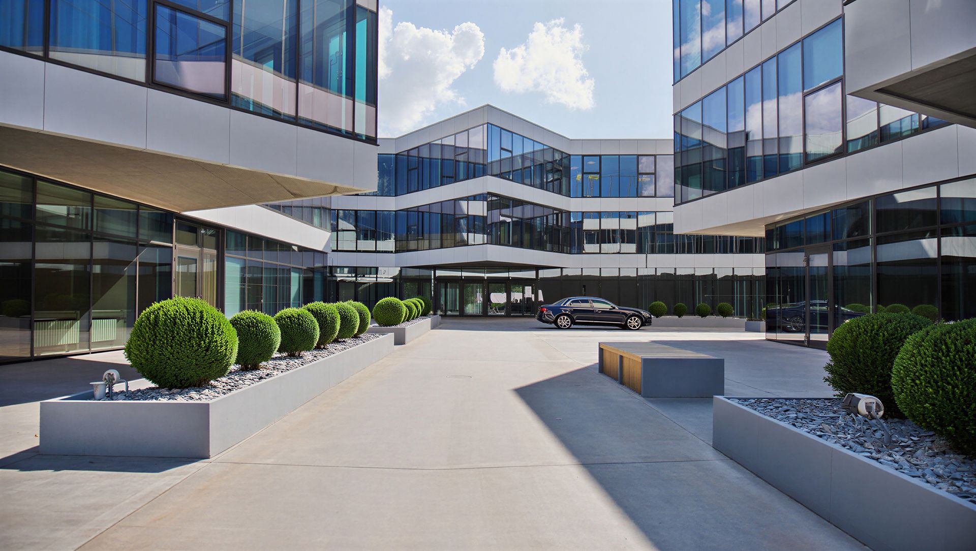 Modern office courtyard with glass buildings, paved walkway, and trimmed shrubs under a bright sky