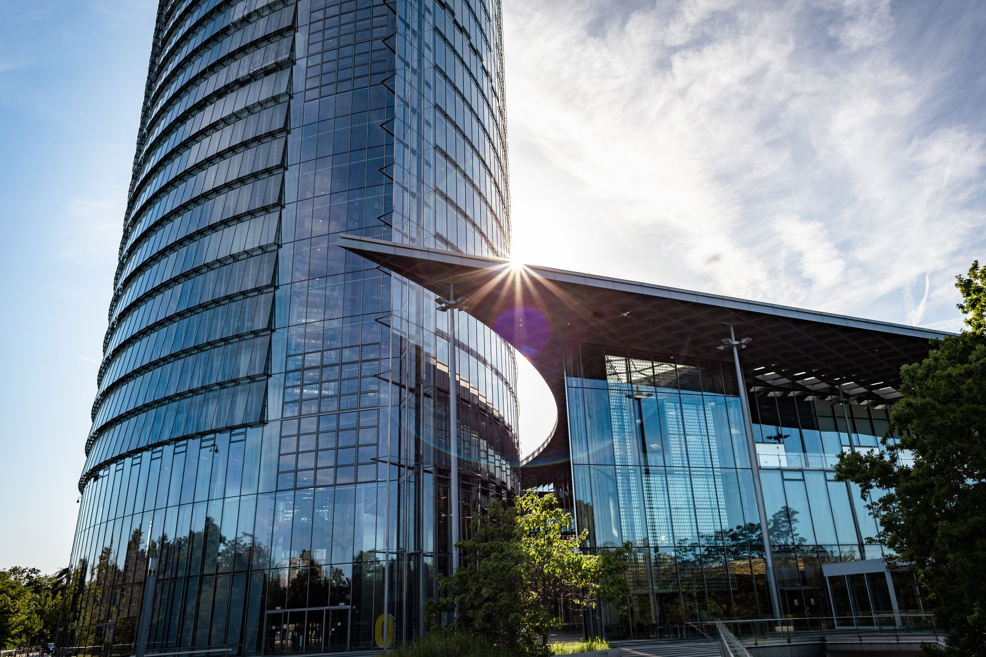 Modern glass office building with a curved tower and sun flare against a blue sky