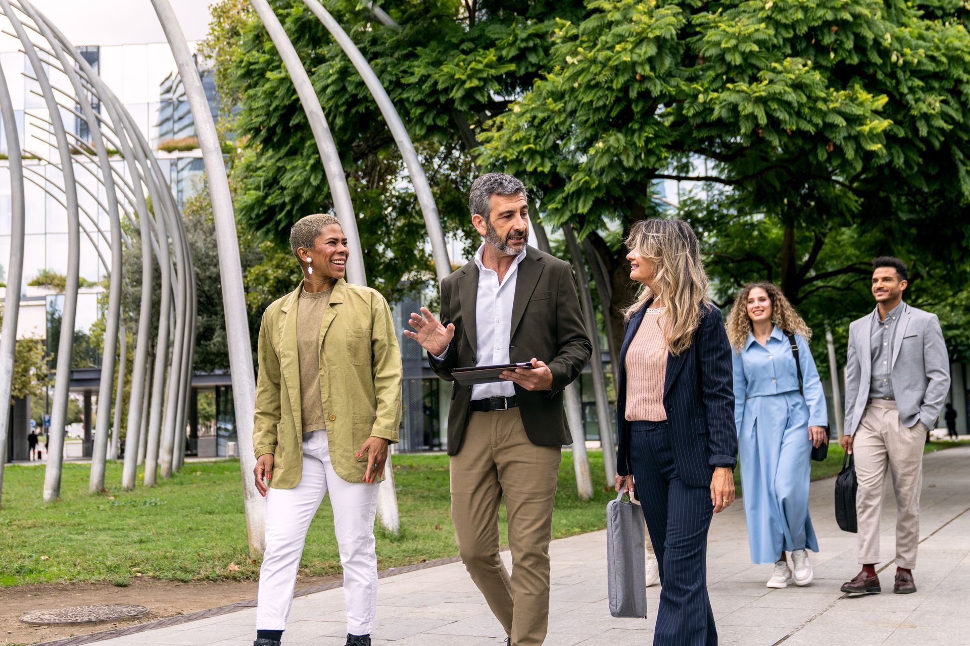 Four people walking and talking on a tree-lined campus path near a modern metal sculpture