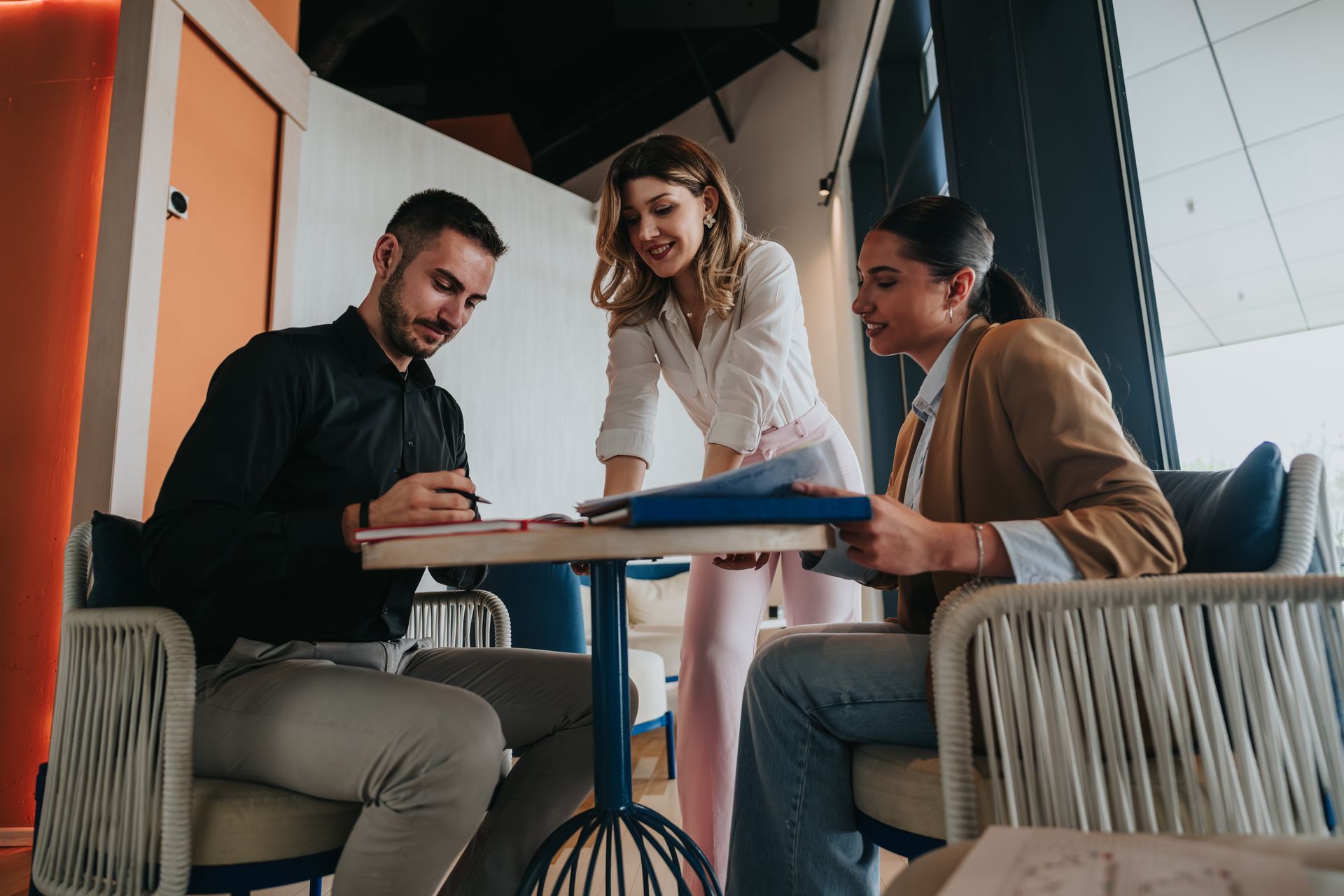 Three coworkers meeting at a table, reviewing documents in a modern office lounge.
