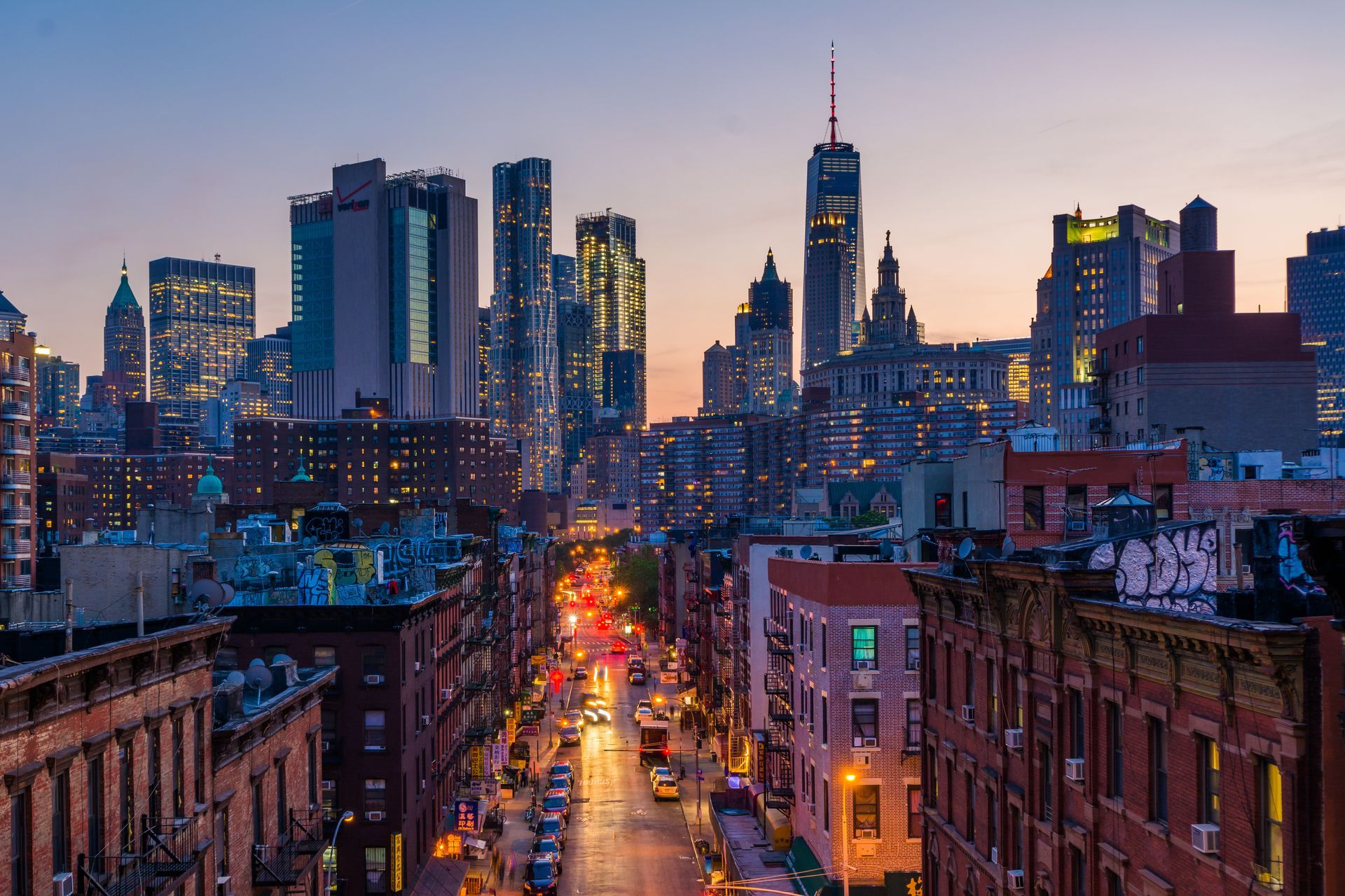 City street at dusk with glowing traffic, flanked by brick buildings and a skyline of tall skyscrapers.