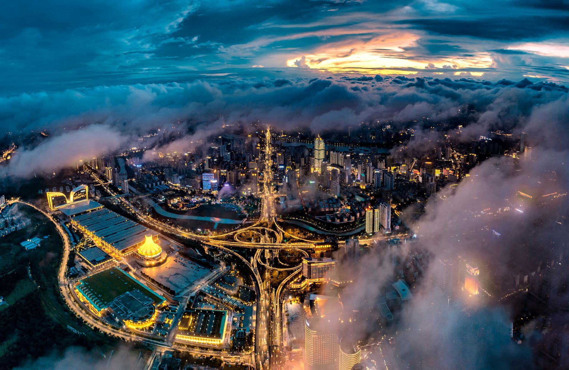 Aerial city skyline at dusk with glowing roads, high-rises, and low clouds over water and mountains