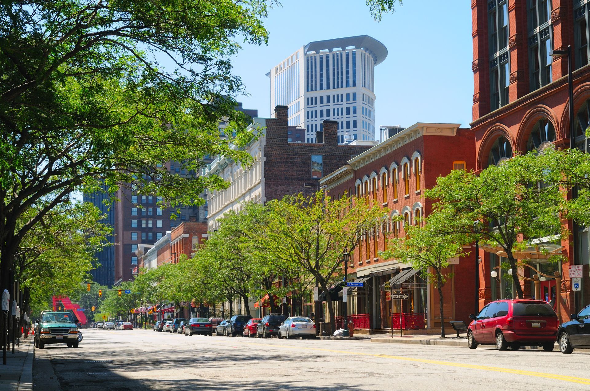 Sunny downtown street with red-brick buildings, parked cars, trees, and a modern high-rise in the background