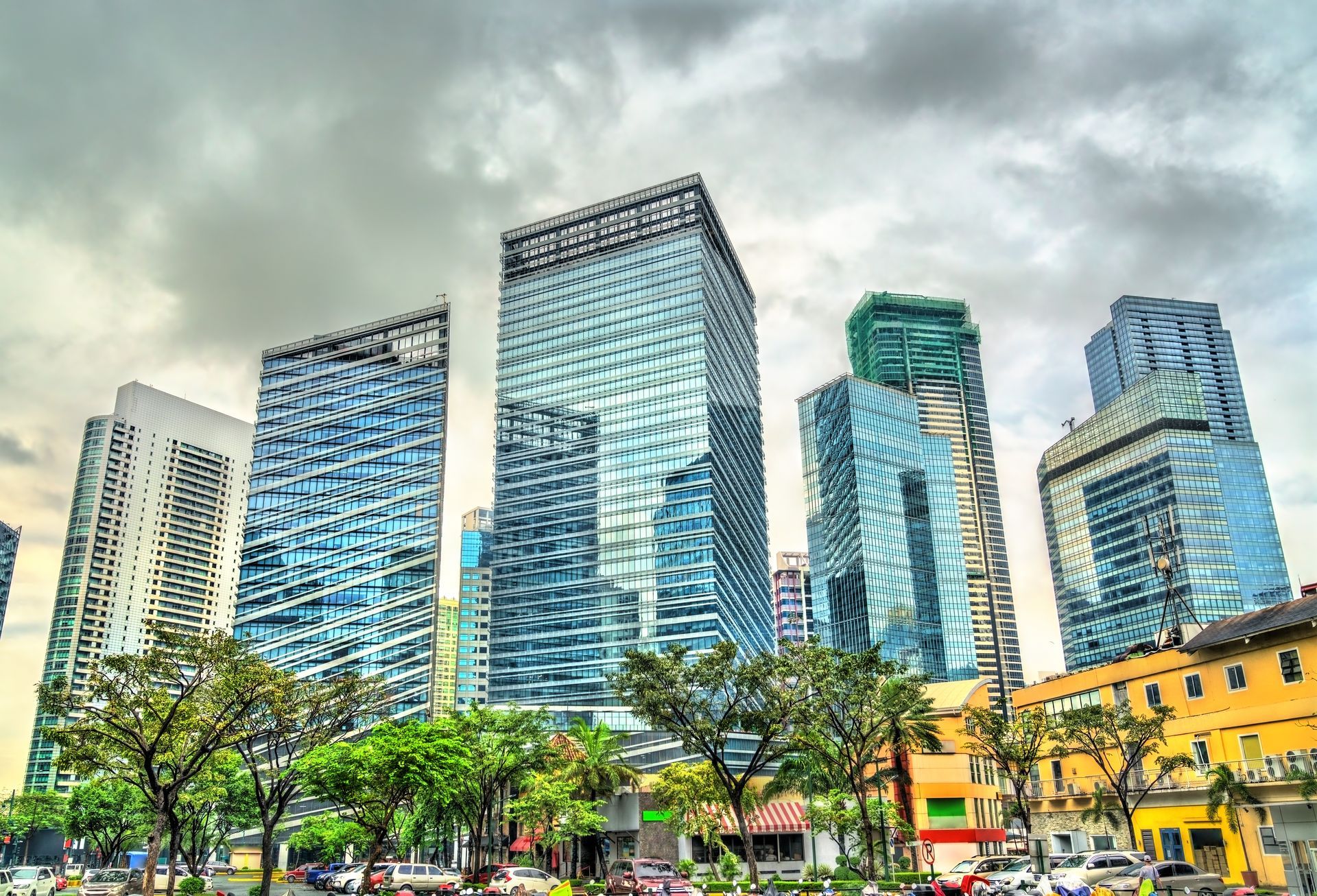 Modern glass skyscrapers above a tree-lined city street on a cloudy day