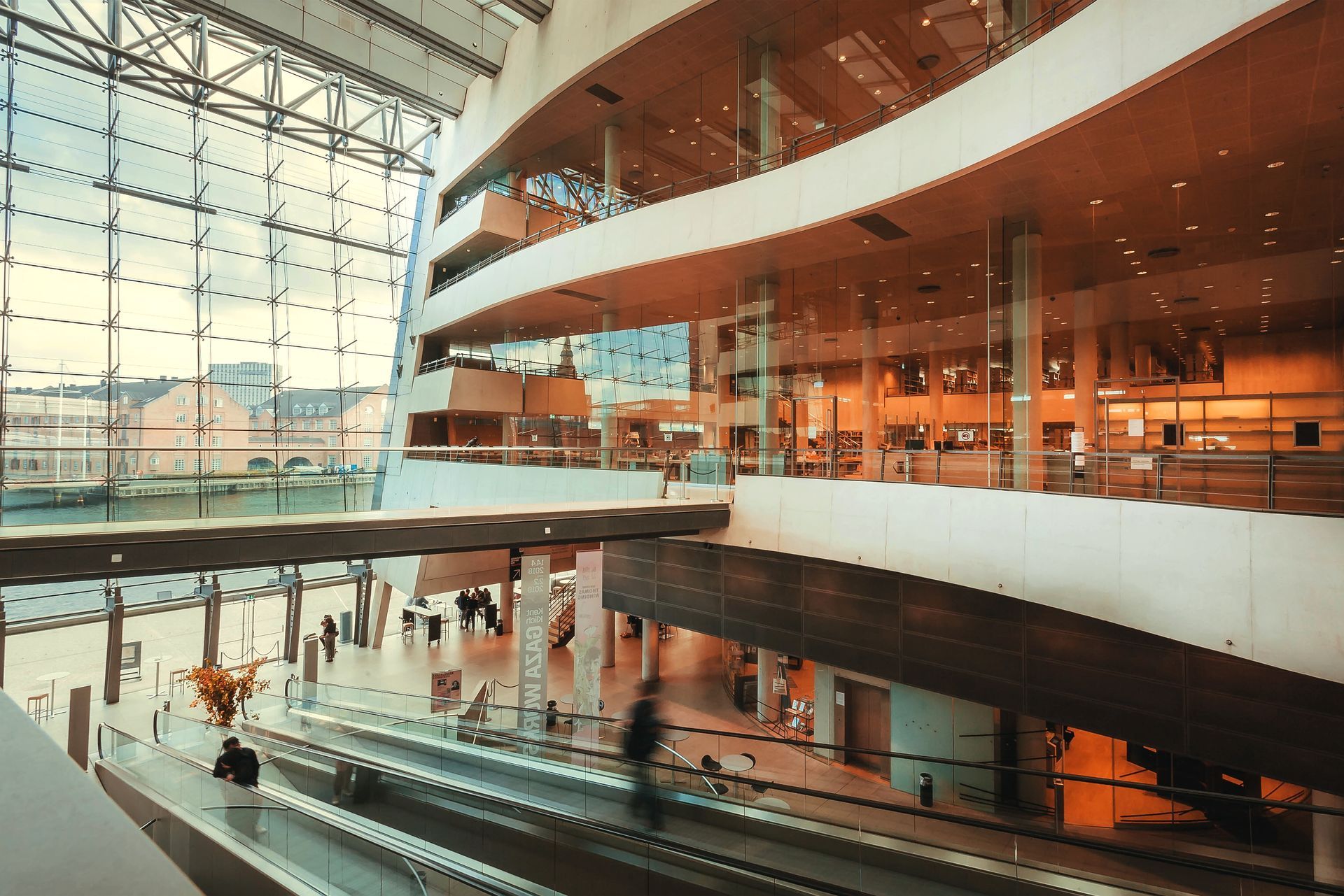 Modern glass atrium with curved balconies, escalators, and people walking inside a bright building