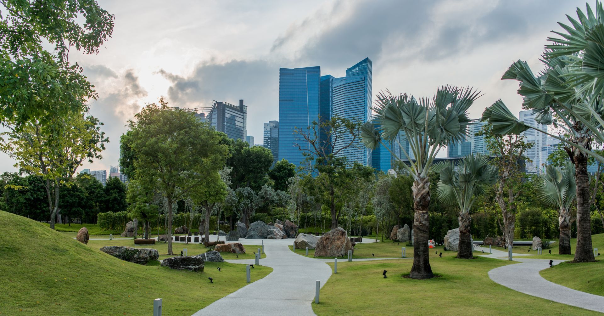 Park with curved pathway, trees, and tall buildings in the background under a cloudy sky.