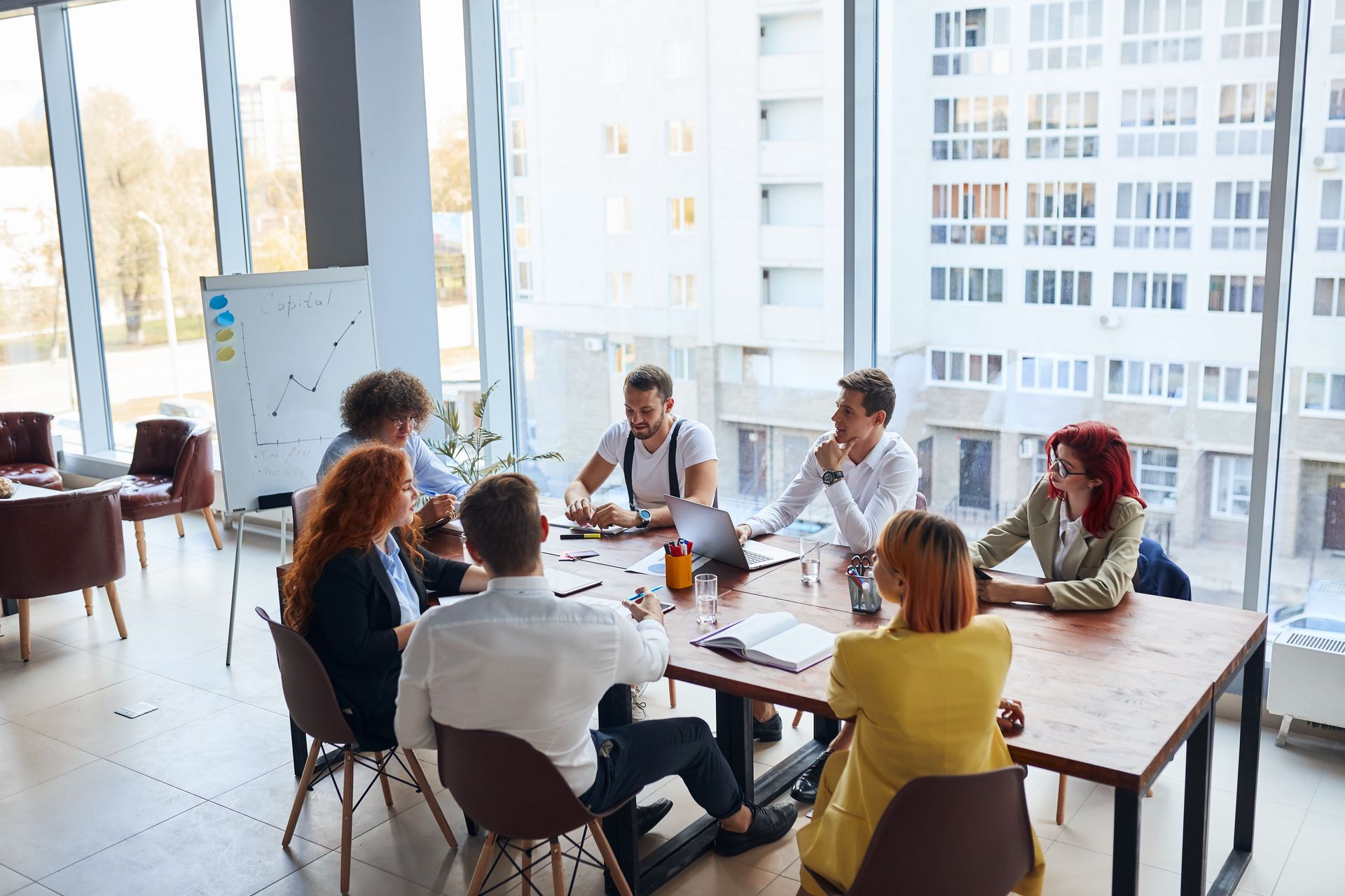 People seated around a conference table in a bright office, meeting and talking with laptops and drinks.