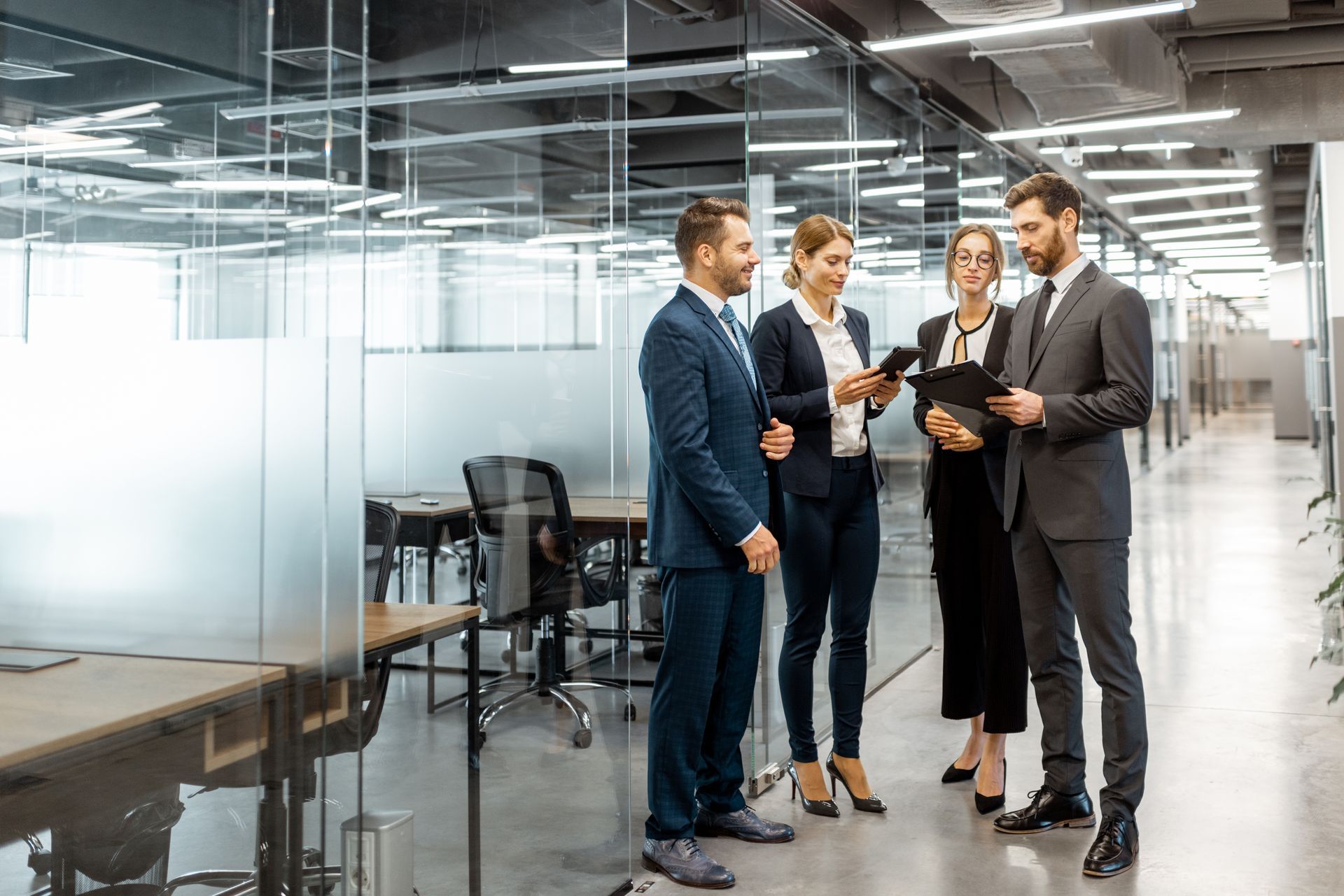 Four business professionals reviewing a tablet in a modern glass-walled office corridor