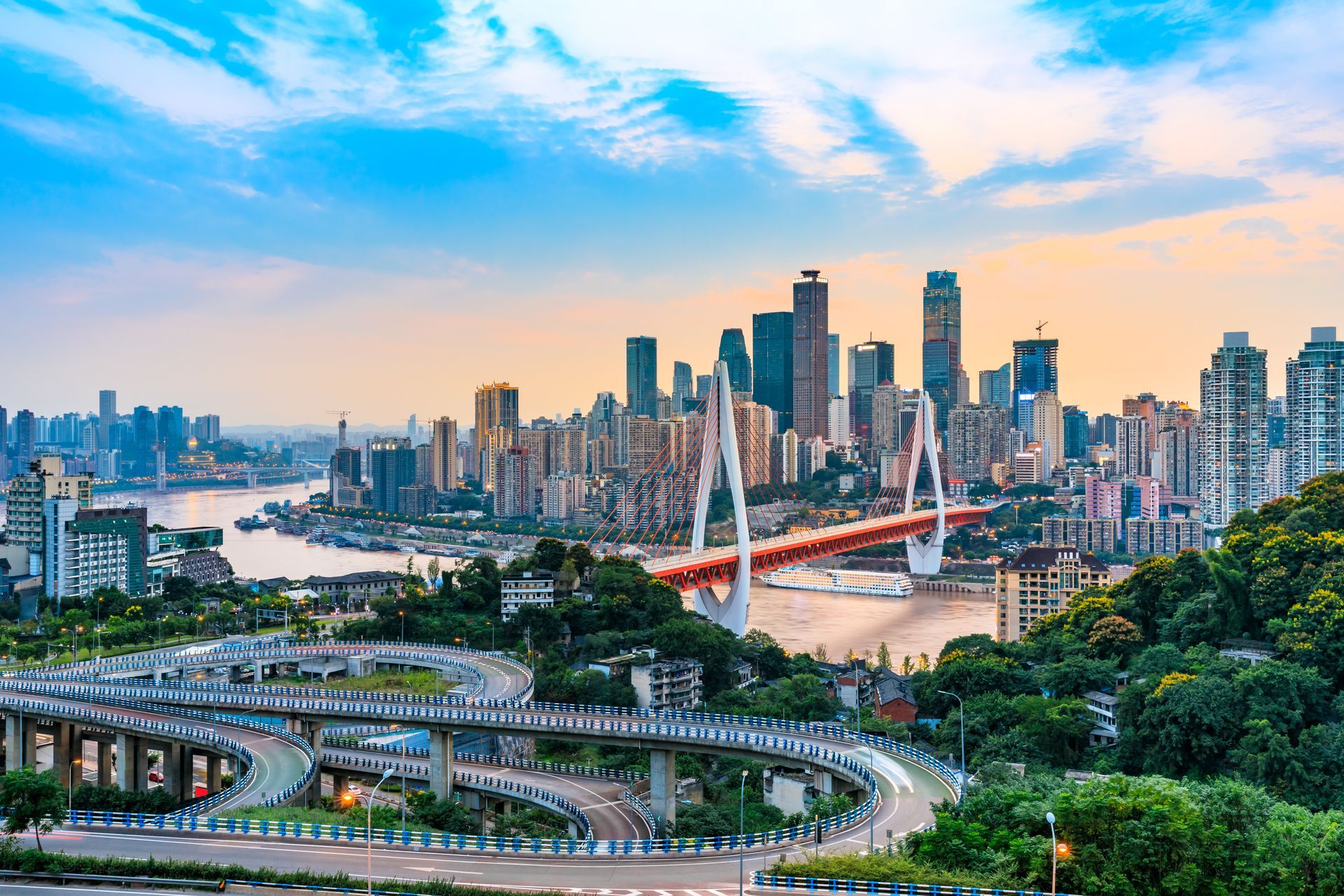 City skyline at sunset with a red bridge, river, and curving highway over lush green hills