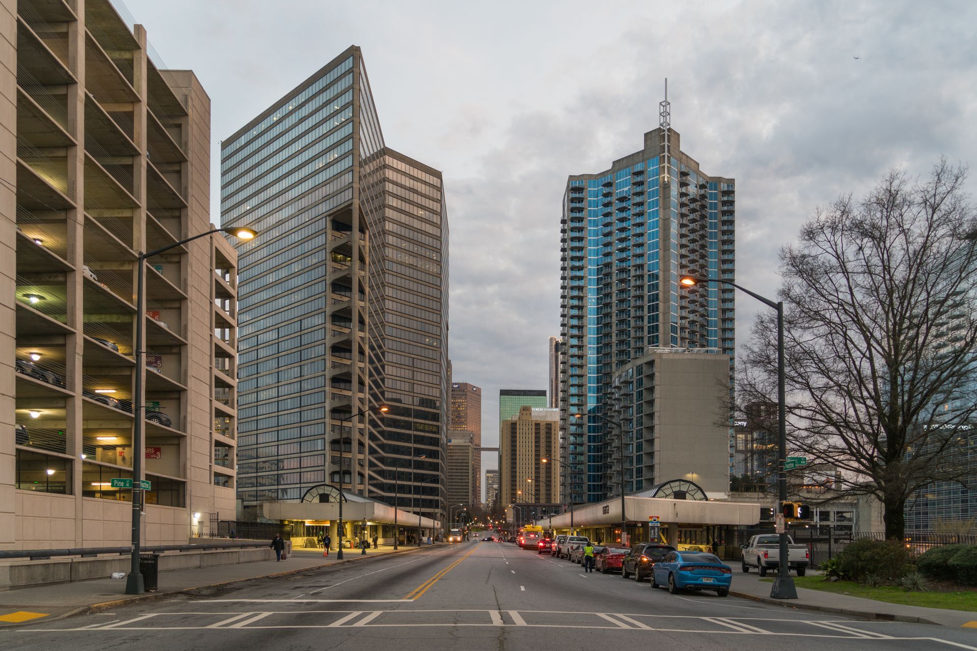 Downtown city street with tall modern buildings and parked cars under a cloudy sky