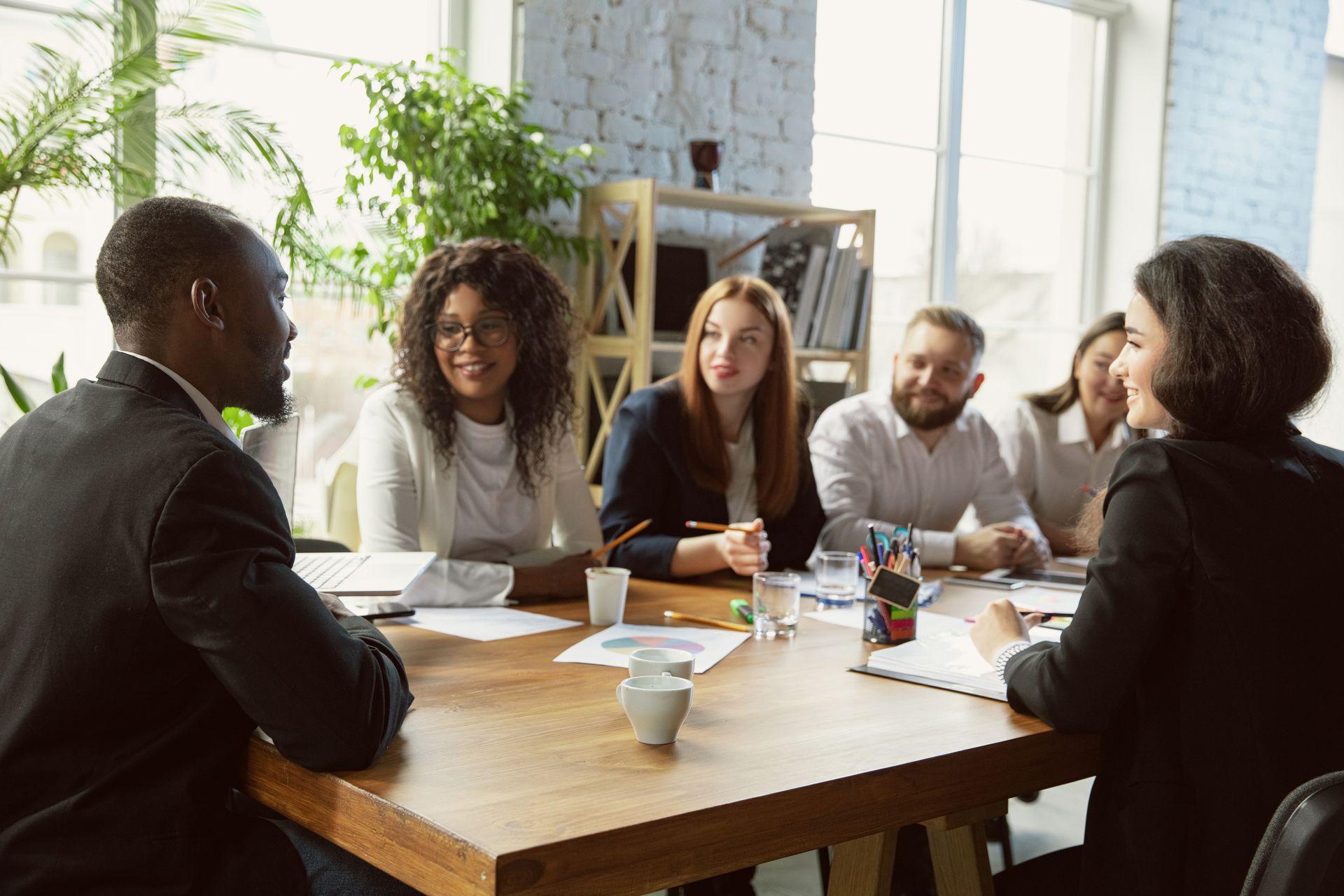 People in a bright office meeting around a wooden table, discussing documents and coffee cups