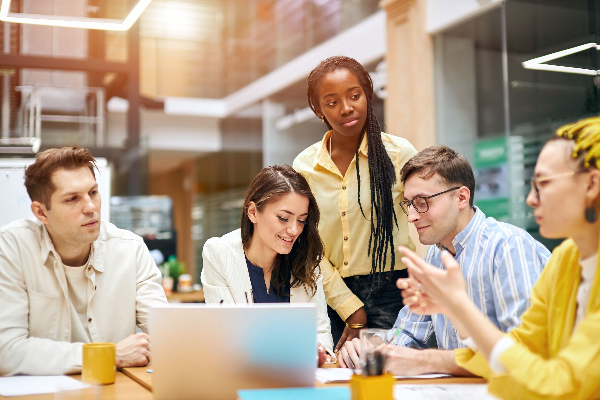 Team meeting around a laptop in a bright office, with yellow mugs and colleagues talking.
