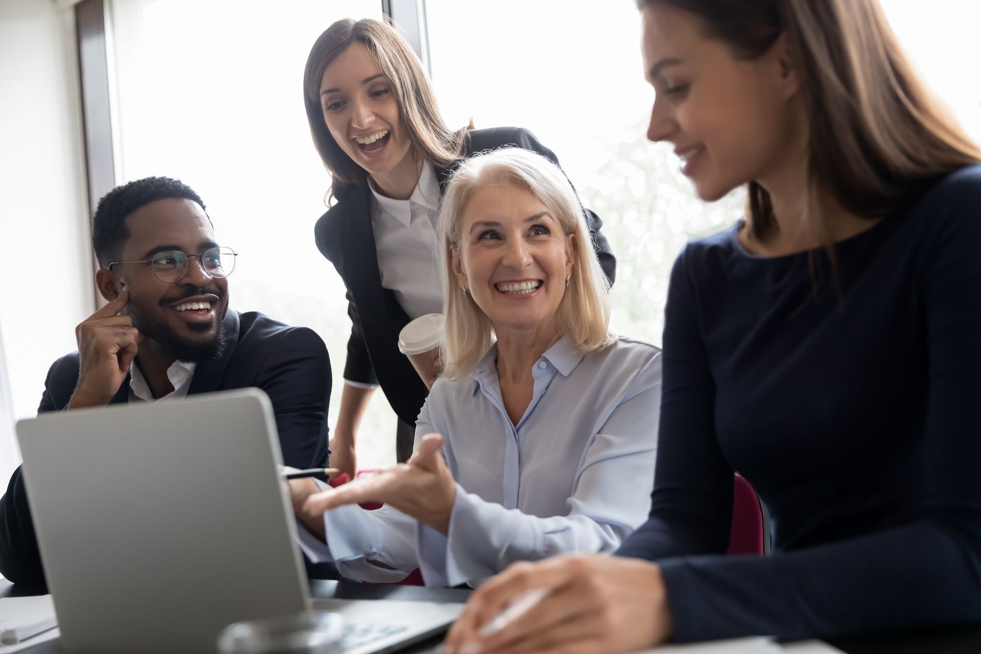 Four coworkers gathered around a laptop in a bright office, smiling and discussing together
