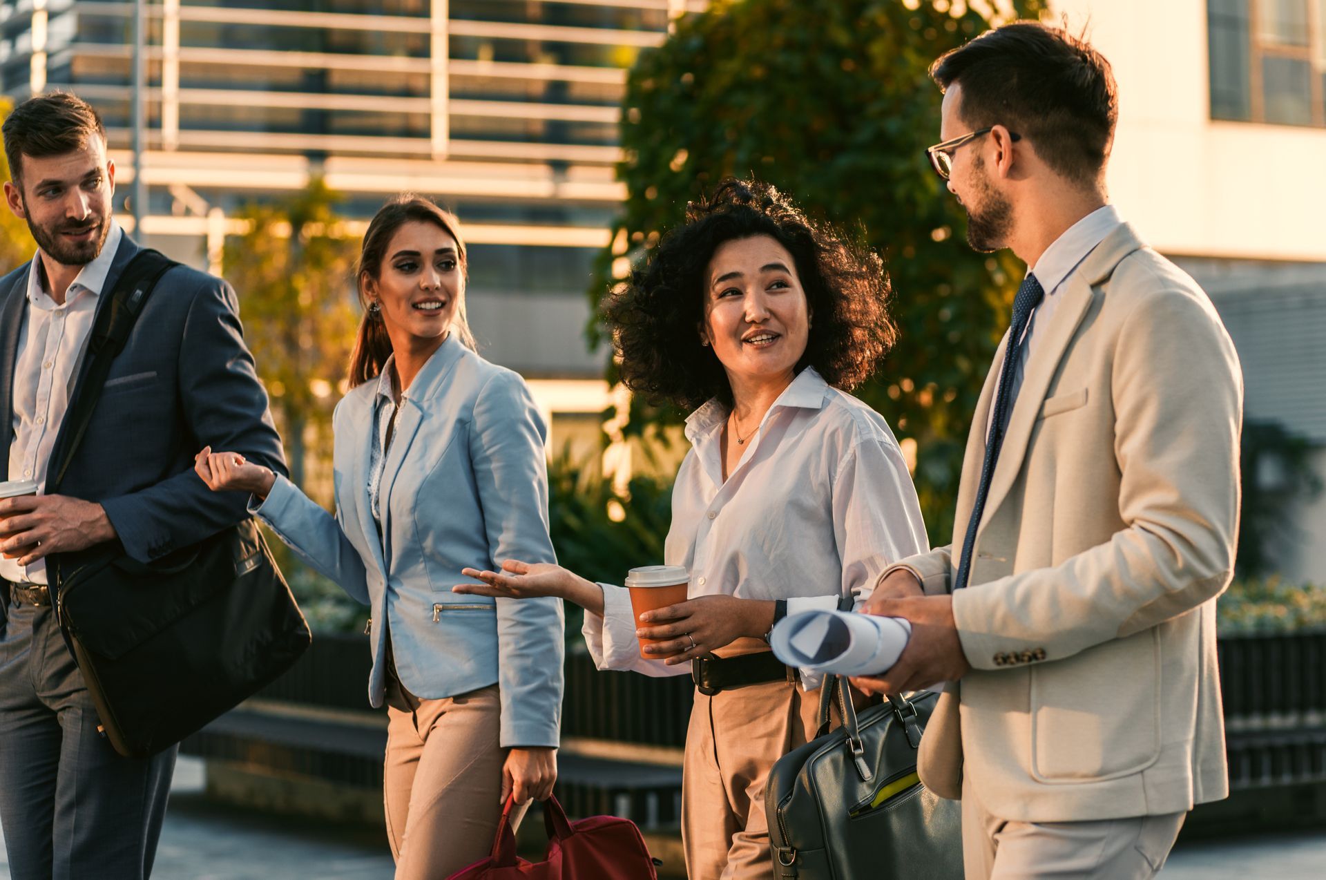 Four business people in formal wear talking outdoors with coffee cups and bags in a city setting