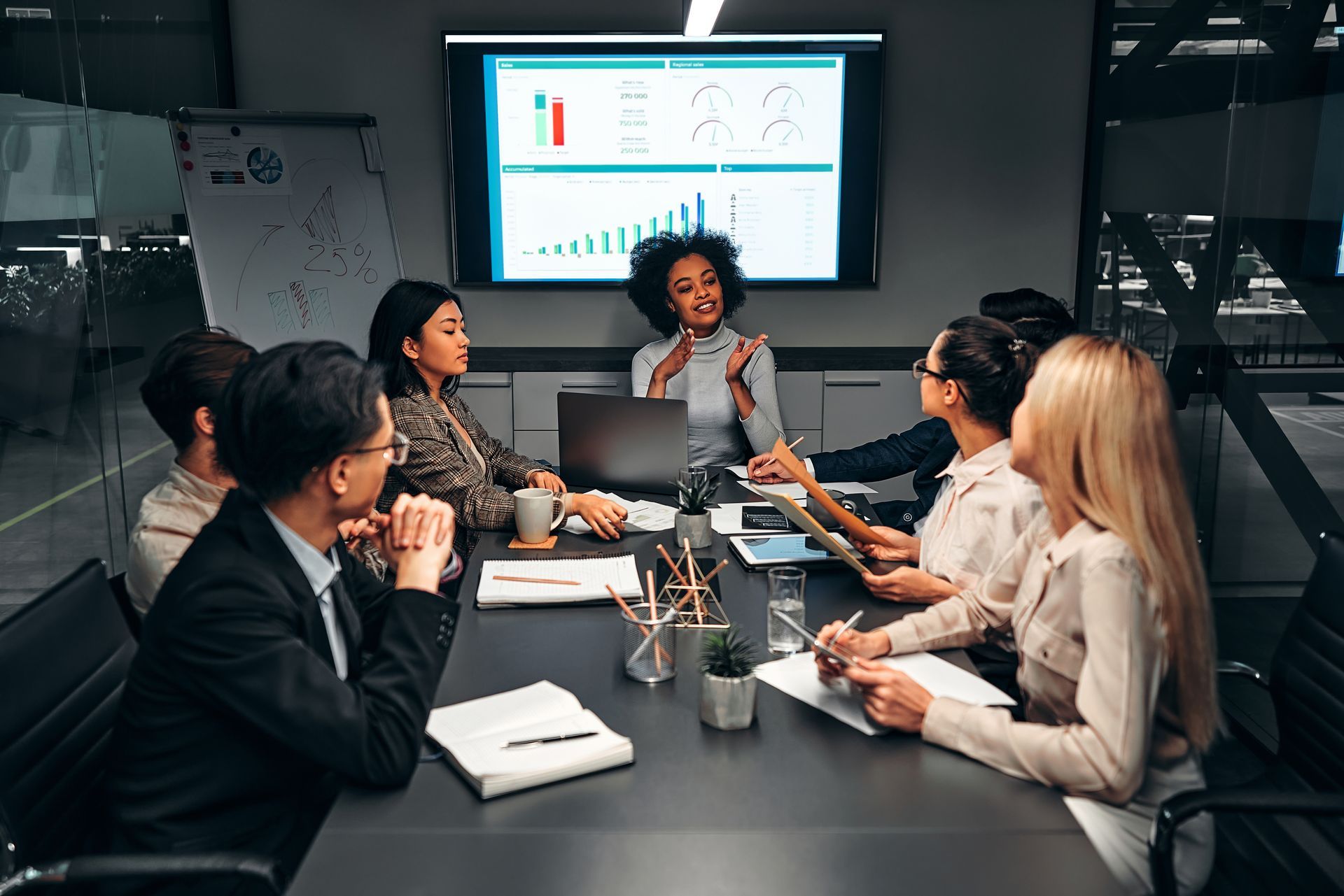 Business meeting around a conference table with a presenter and a screen showing charts and graphs