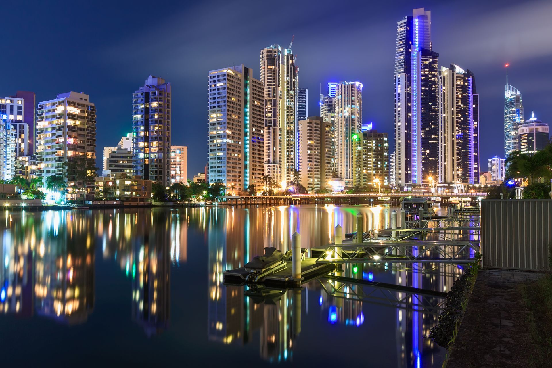City skyline at night reflected on calm water, with brightly lit high-rise buildings and colorful lights.